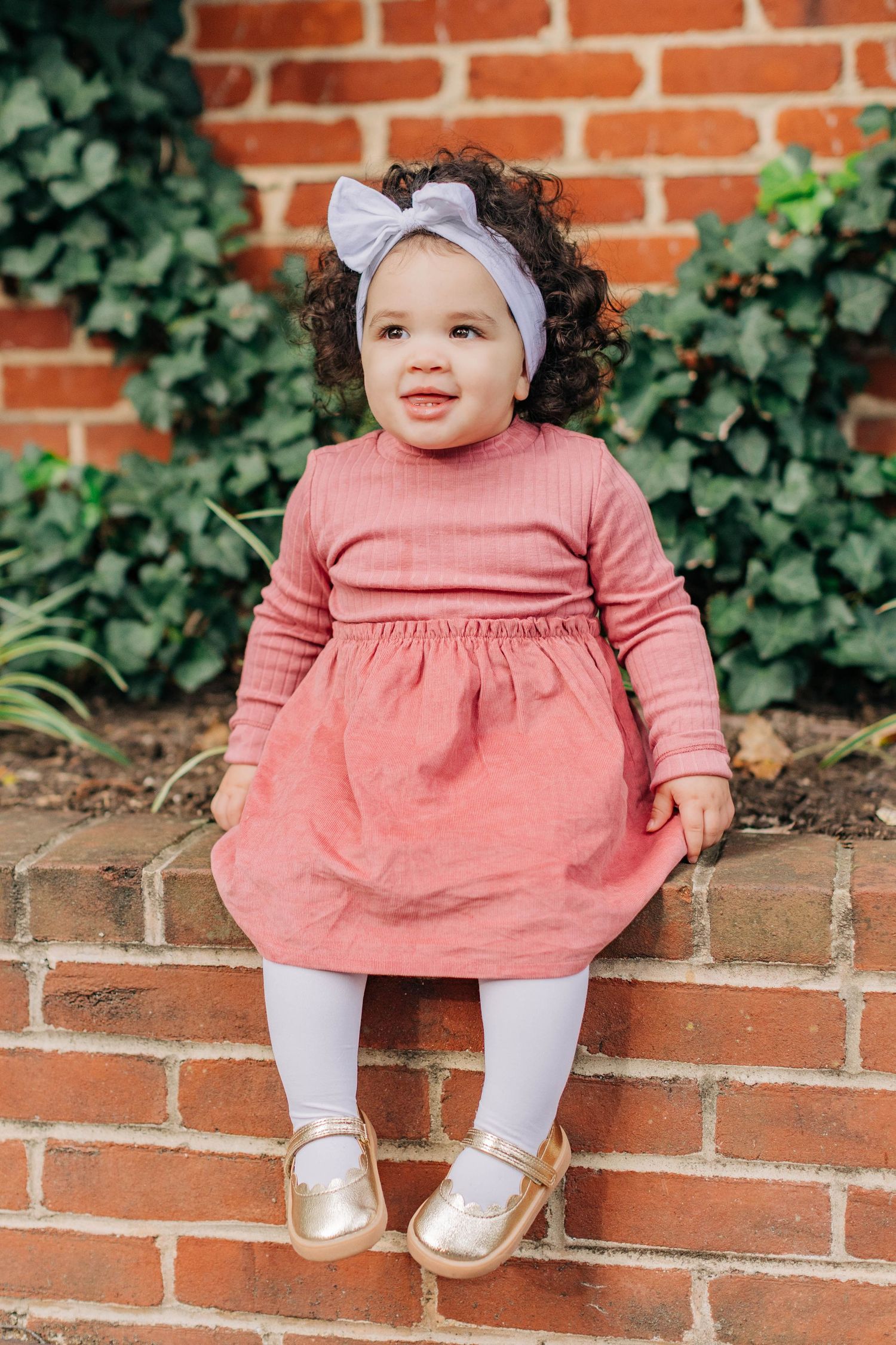 Toddler girl in pink dress, white tights, gold shoes, and gray bow headband sitting on brick wall outdoors.