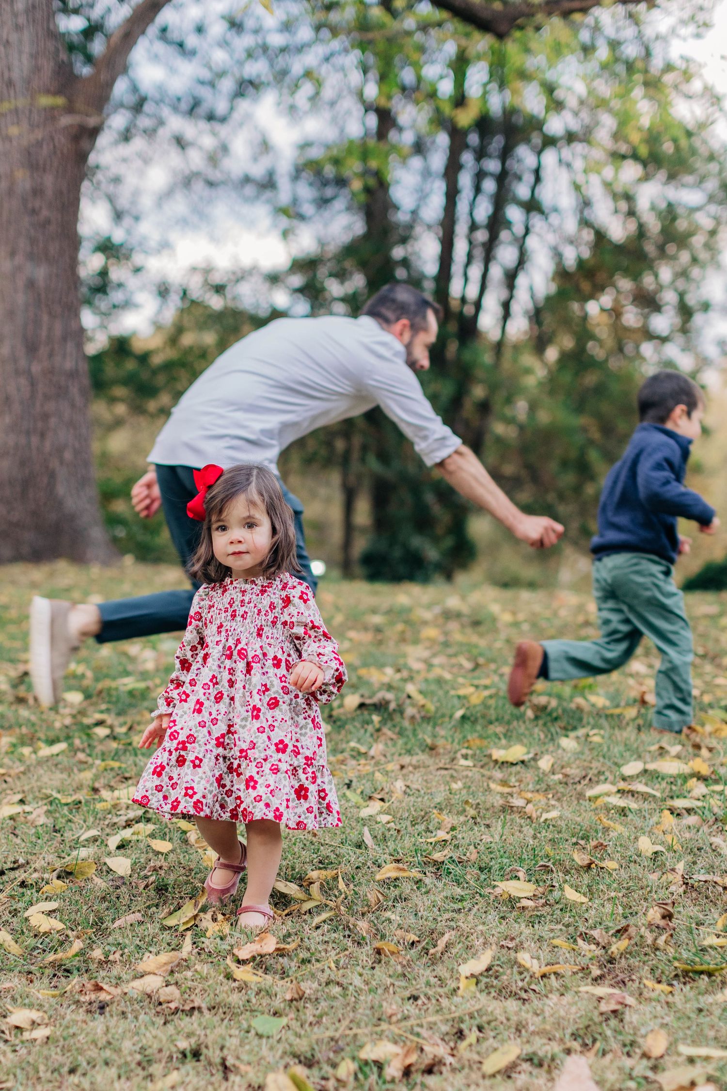 A toddler girl in a floral dress stands in a park while her father plays with another child among autumn leaves.
