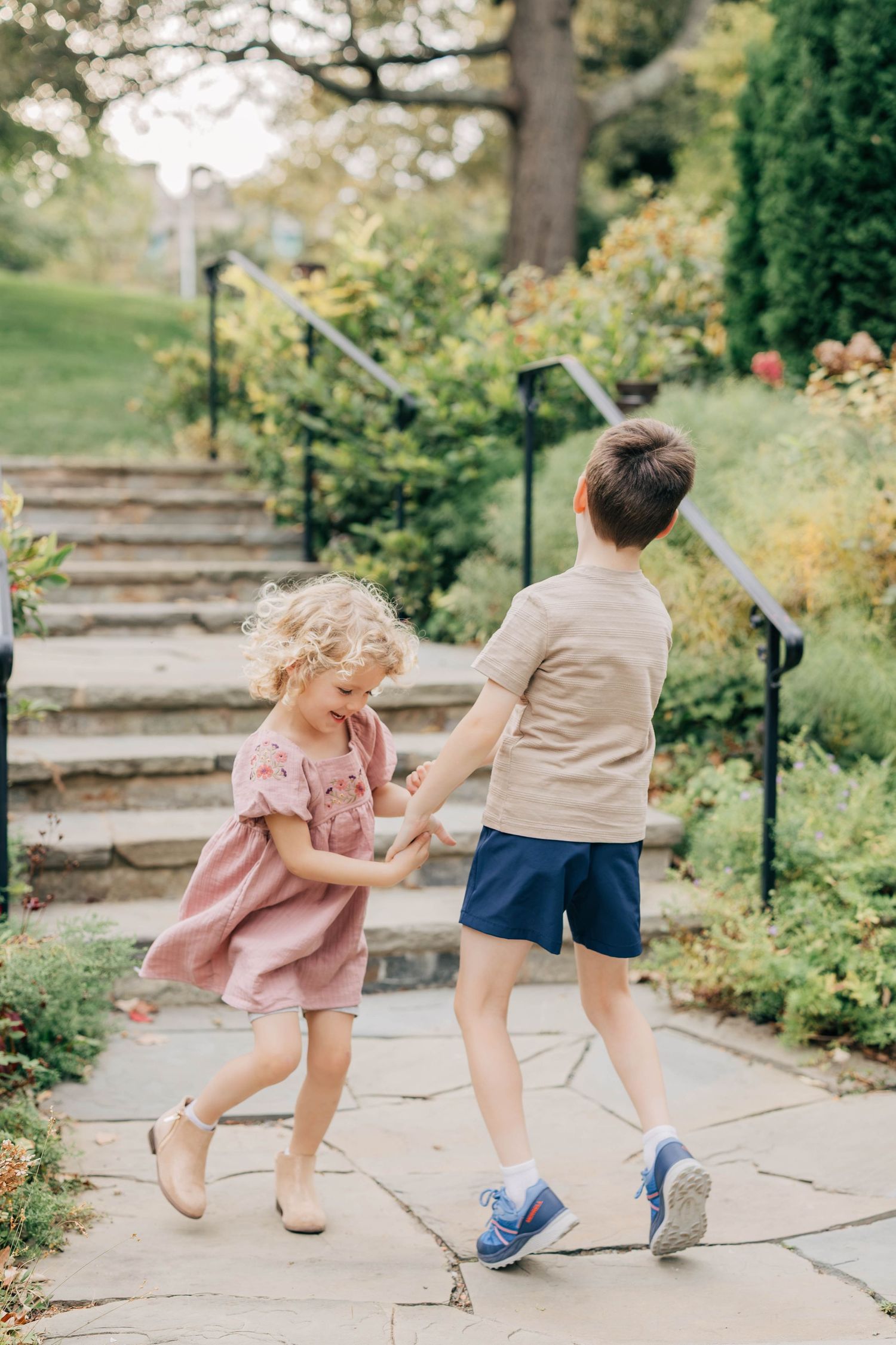 Two young children hold hands and play on a stone path beside garden steps, the girl in a pink dress and the boy in blue shorts.