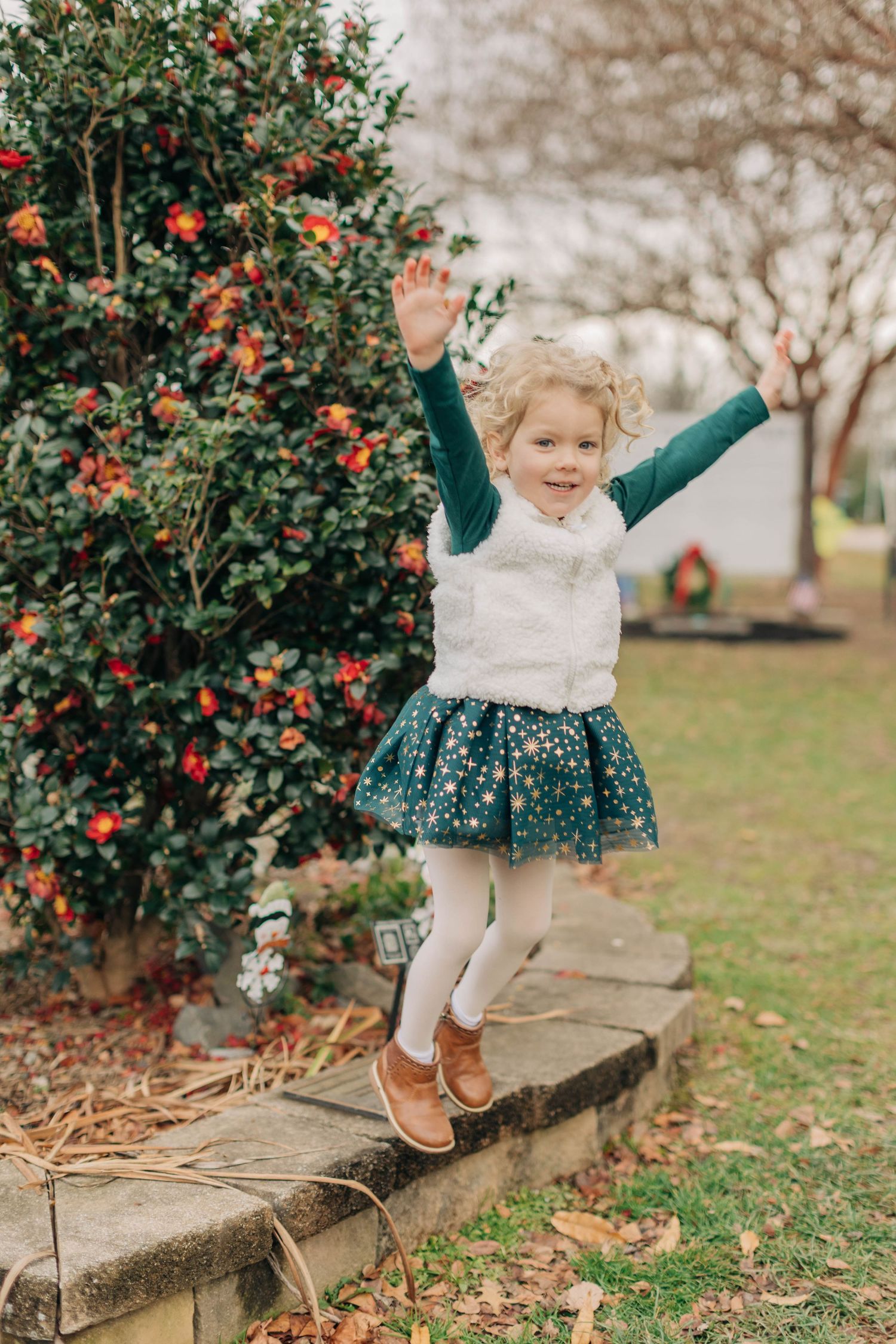 A joyful toddler girl in a green star skirt and white vest jumps with arms raised near a flowering bush outdoors.