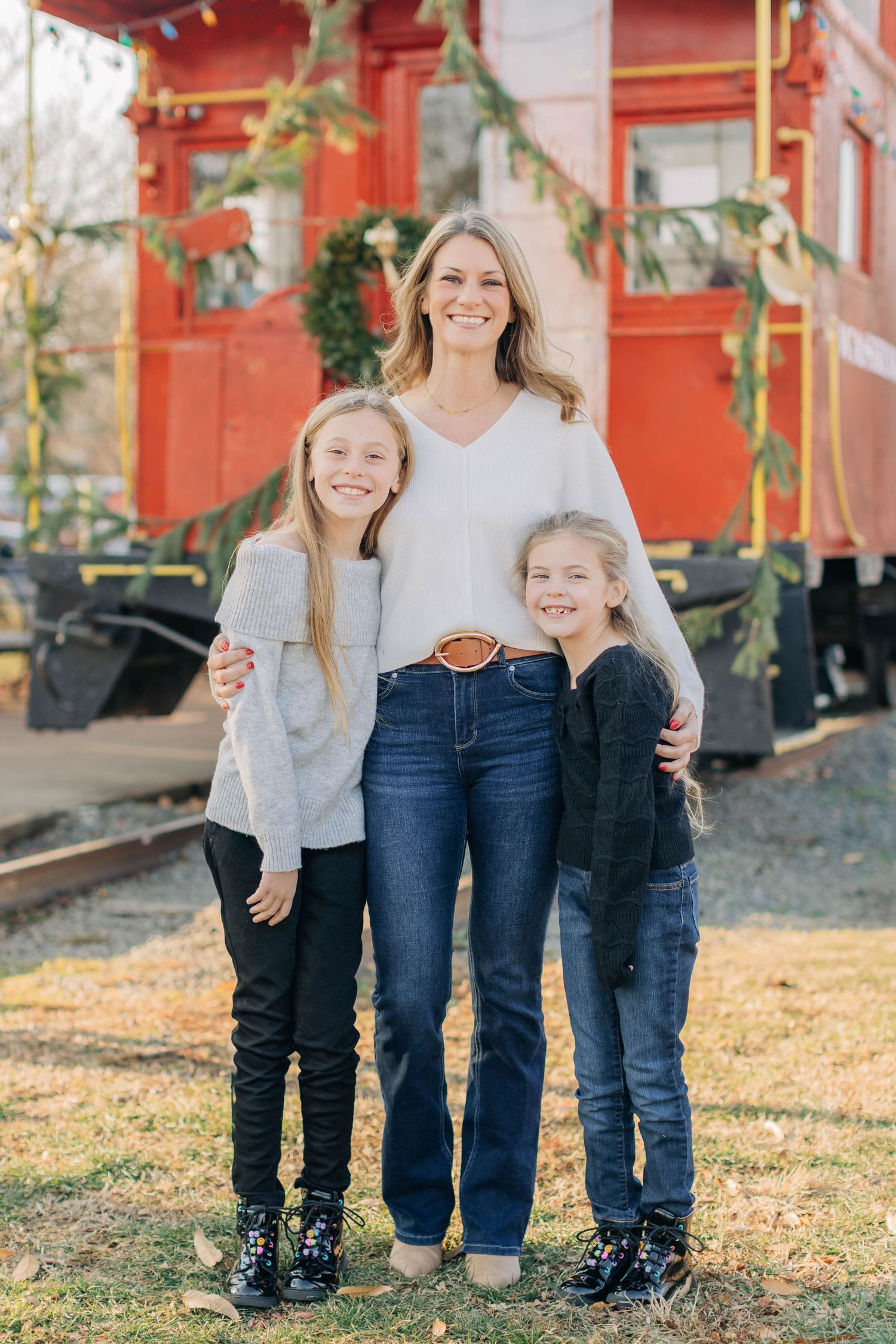 A smiling woman poses with two young girls in front of a red train caboose decorated for Christmas.