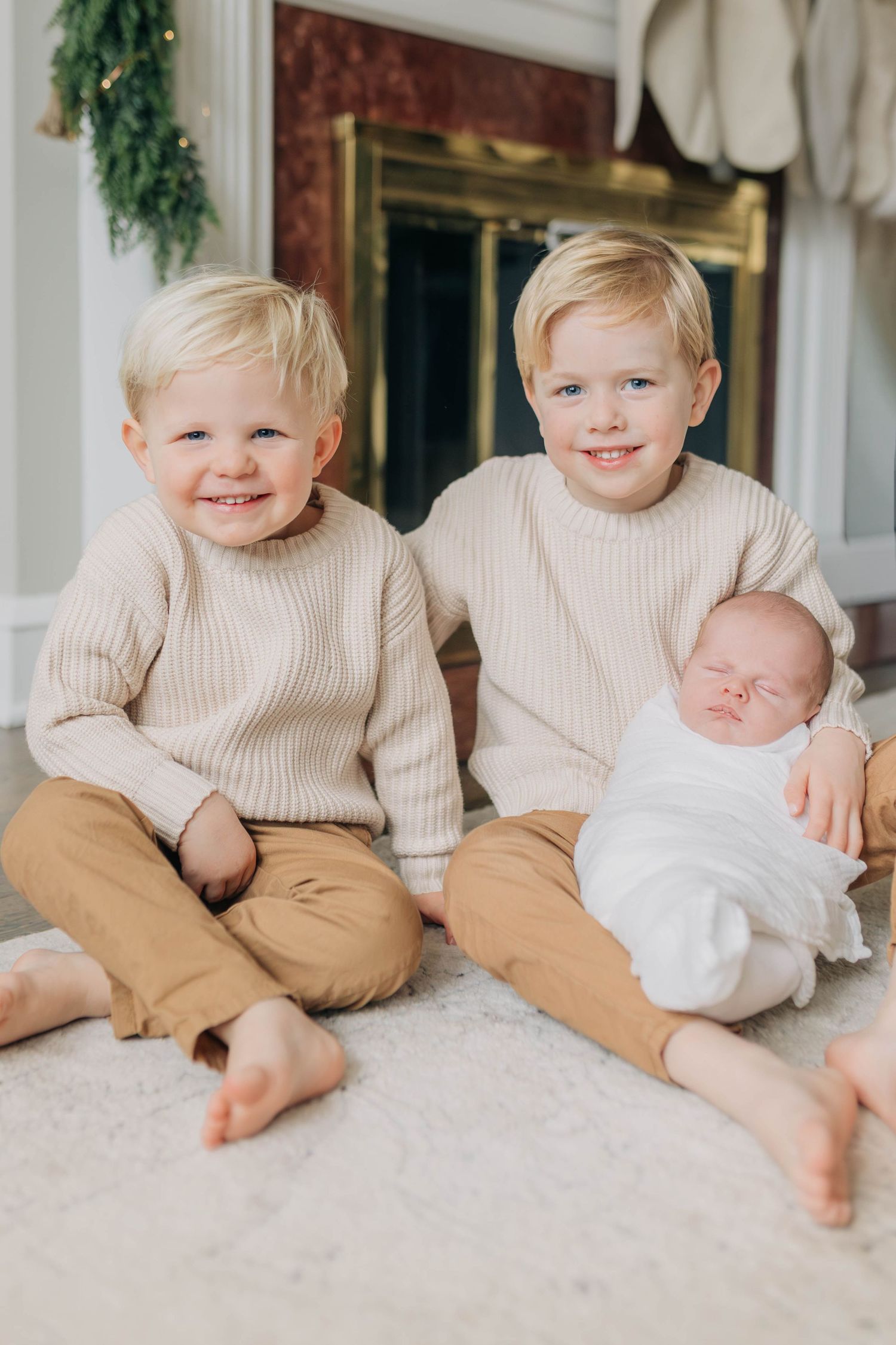 Two children in matching beige sweaters sit with a sleeping baby on a carpet.