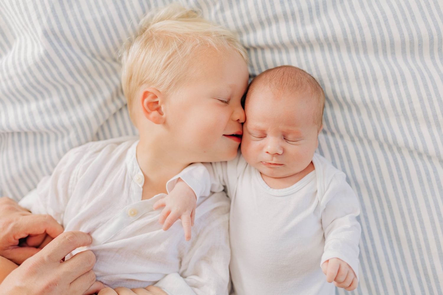 Two children in white clothing share an affectionate moment on striped bedding.