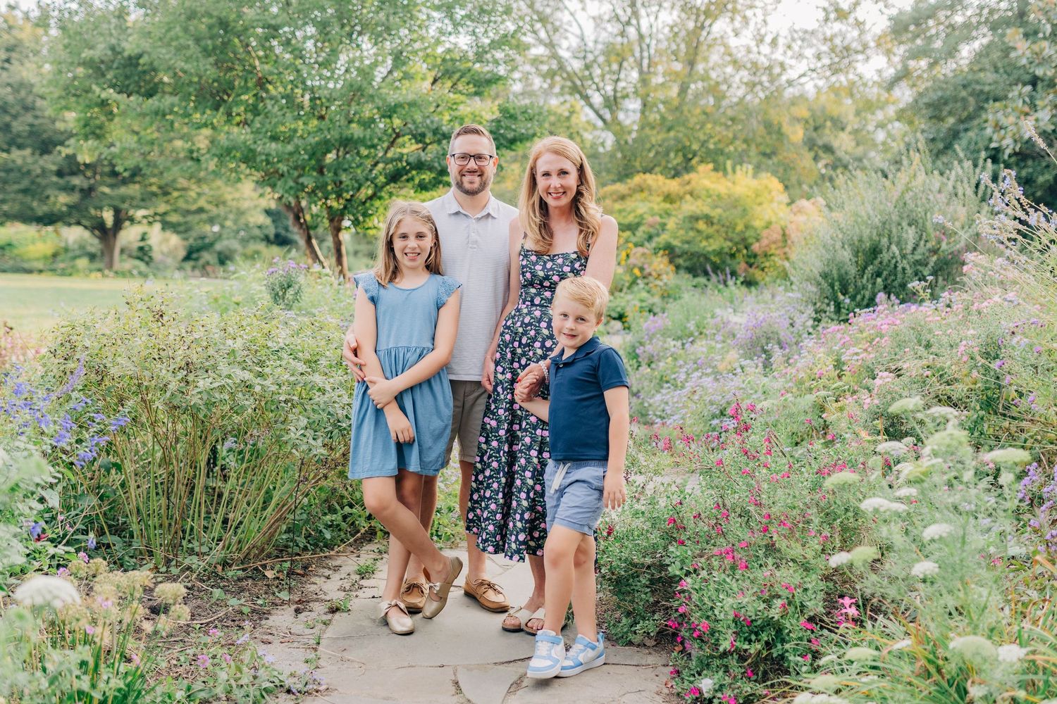 A family of four poses on a garden path surrounded by colorful summer flowers and greenery.