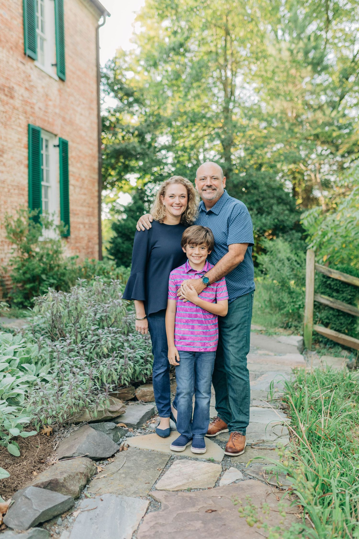 A family poses together on stone steps surrounded by greenery and a brick building with green shutters.