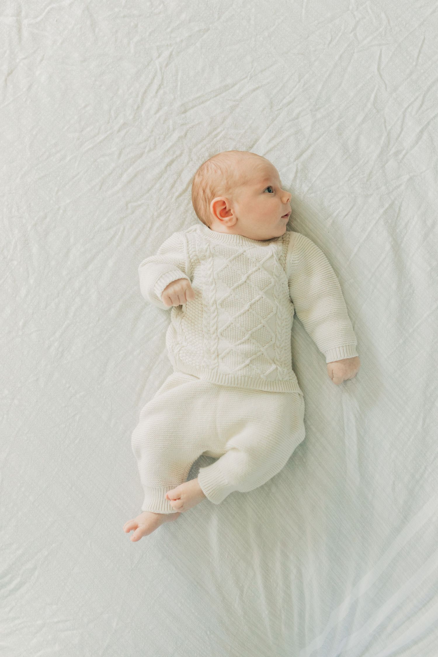 Infant in cream colored knit outfit resting on white bedding.