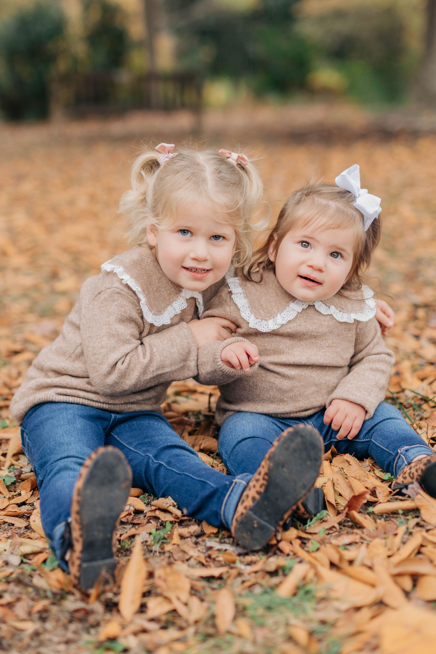 Two young sisters in matching beige sweaters and jeans sit together on autumn leaves in a park.