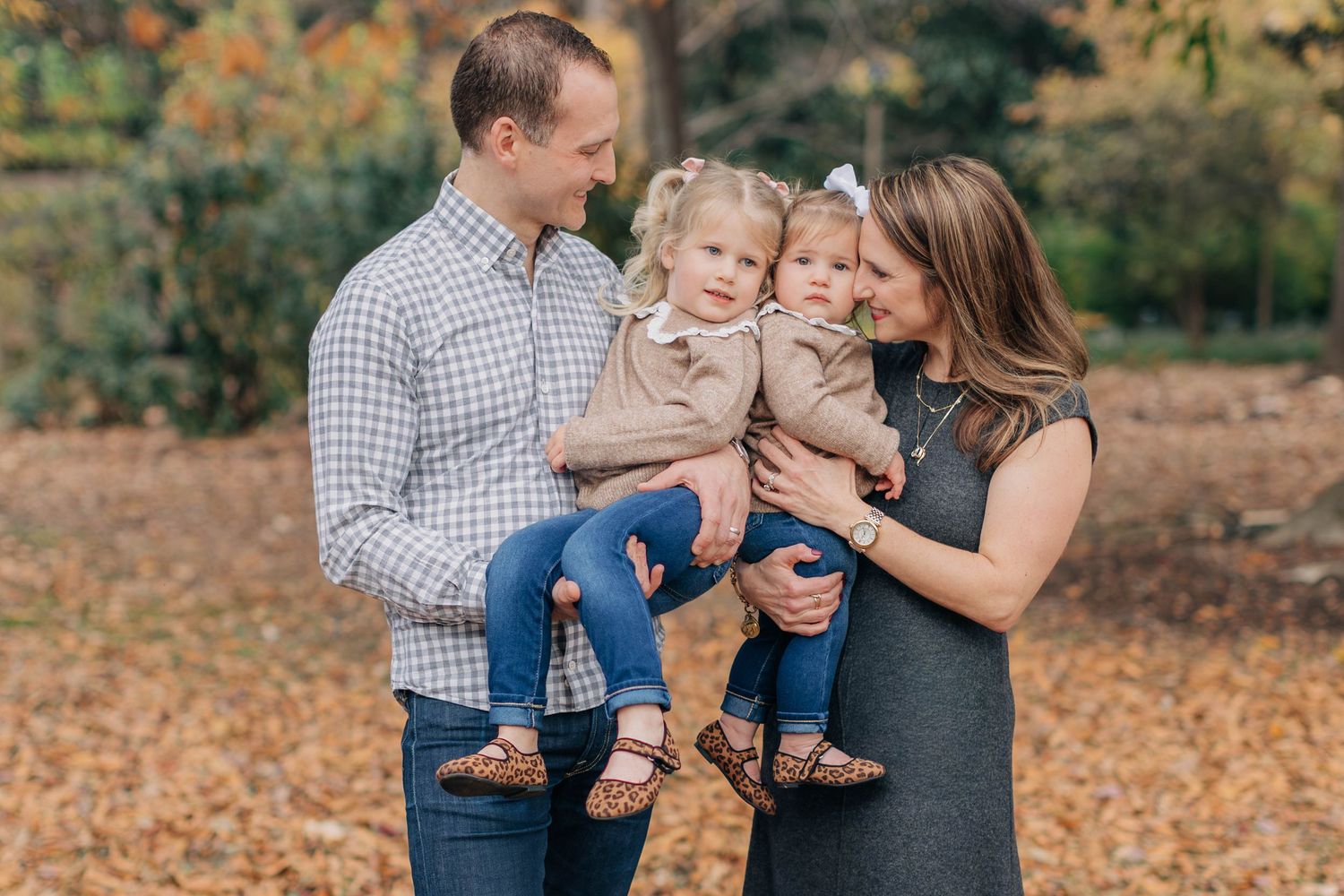Family of four poses outdoors in autumn leaves, parents holding two toddler girls in a warm fall family portrait.