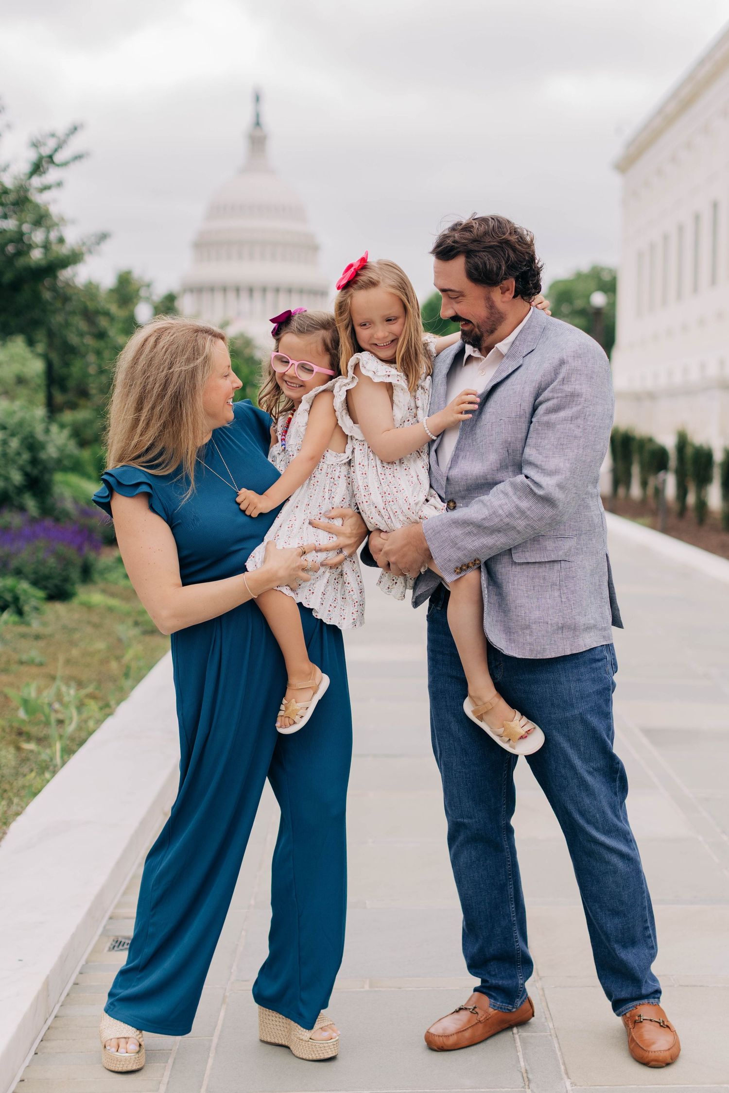 Family of four posing near the U.S. Capitol building, parents holding two young girls in matching outfits on a cloudy day.