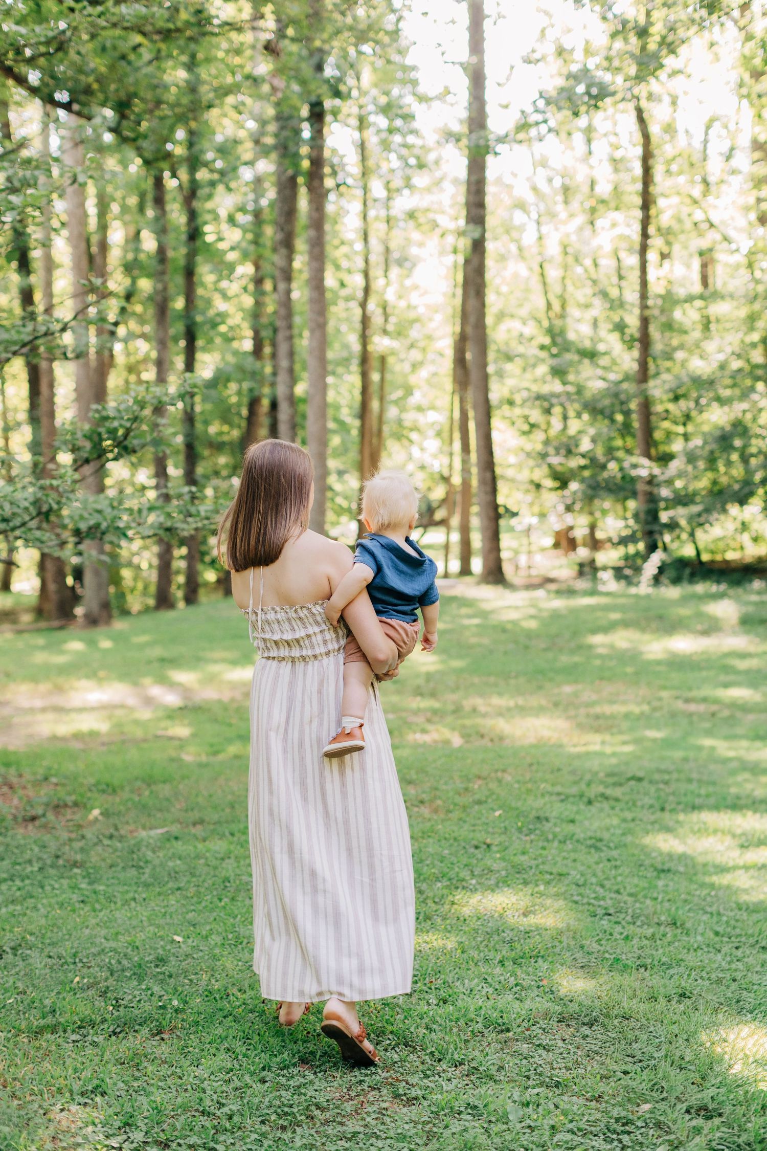 Mother in striped maxi dress holding baby in lush green forest park setting, viewed from behind.