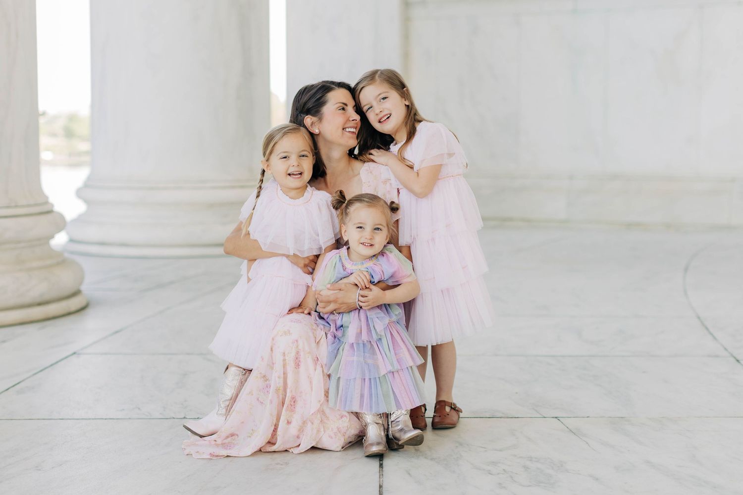 A mother in a pink dress kneels with her three young daughters in matching pink dresses at a marble monument.