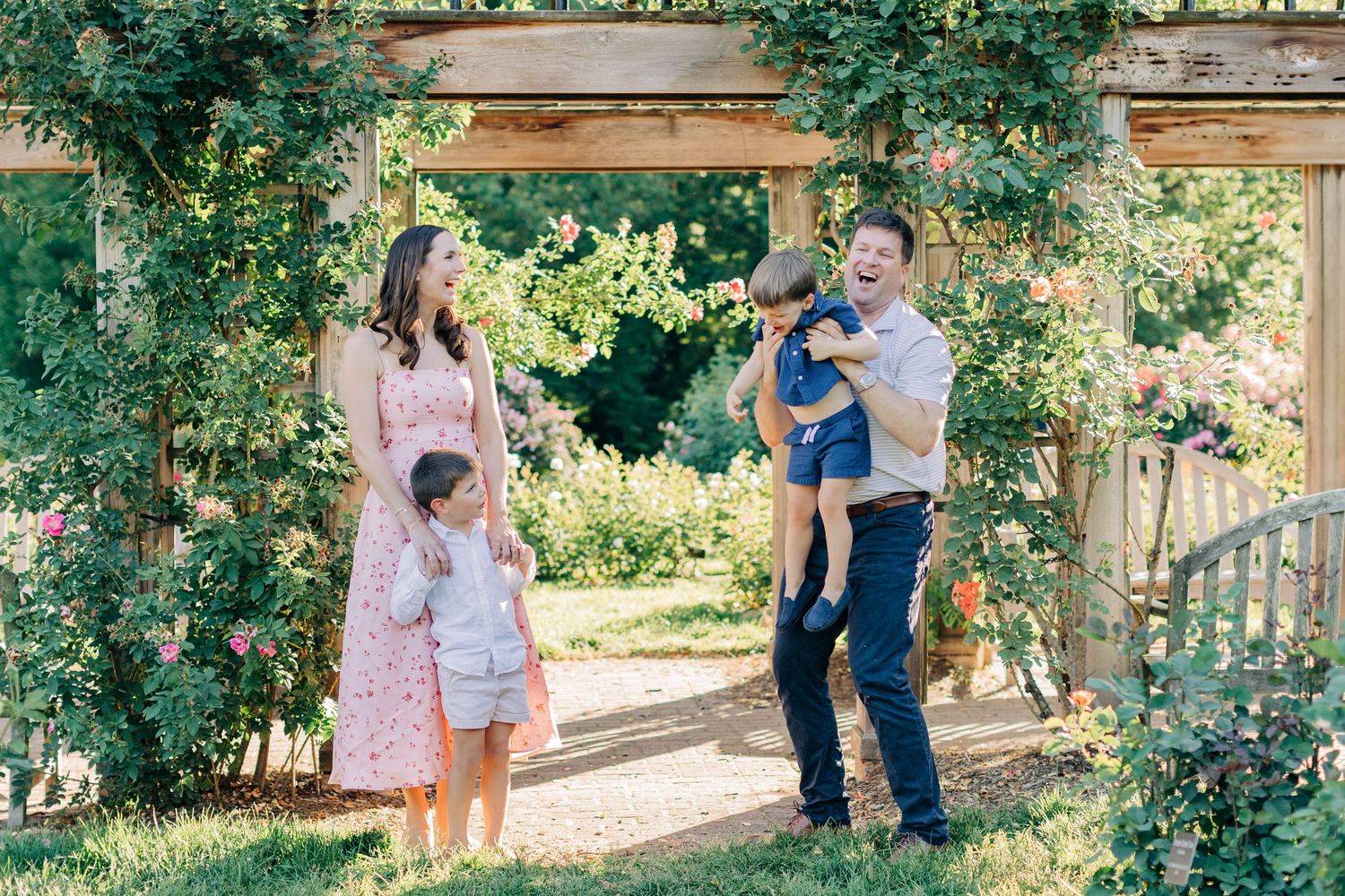 Joyful family of four laughing together under a floral garden arbor on a sunny day.