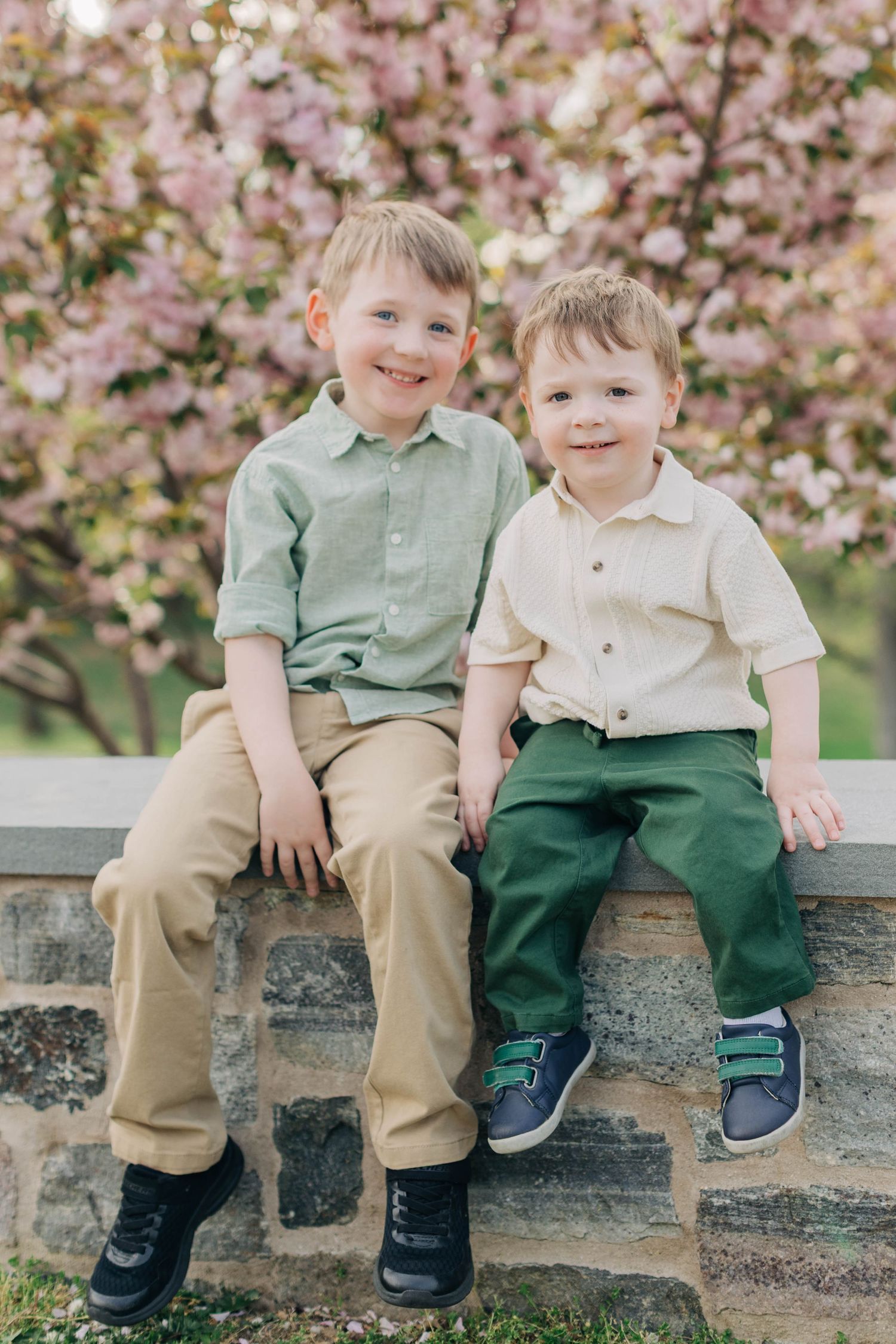 Two smiling boys sit on a stone wall in front of pink cherry blossom trees, wearing green and cream button-up shirts.