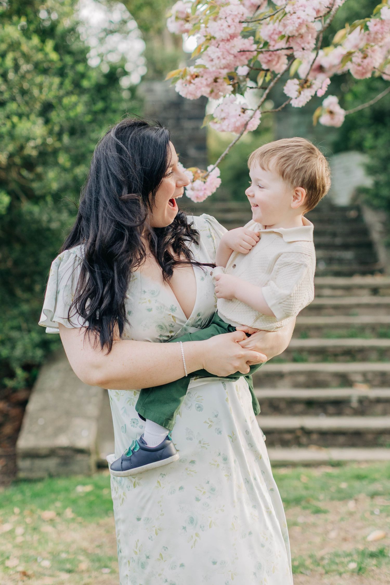 A mother with dark hair in a floral dress holds her smiling toddler boy under pink cherry blossoms in a garden.