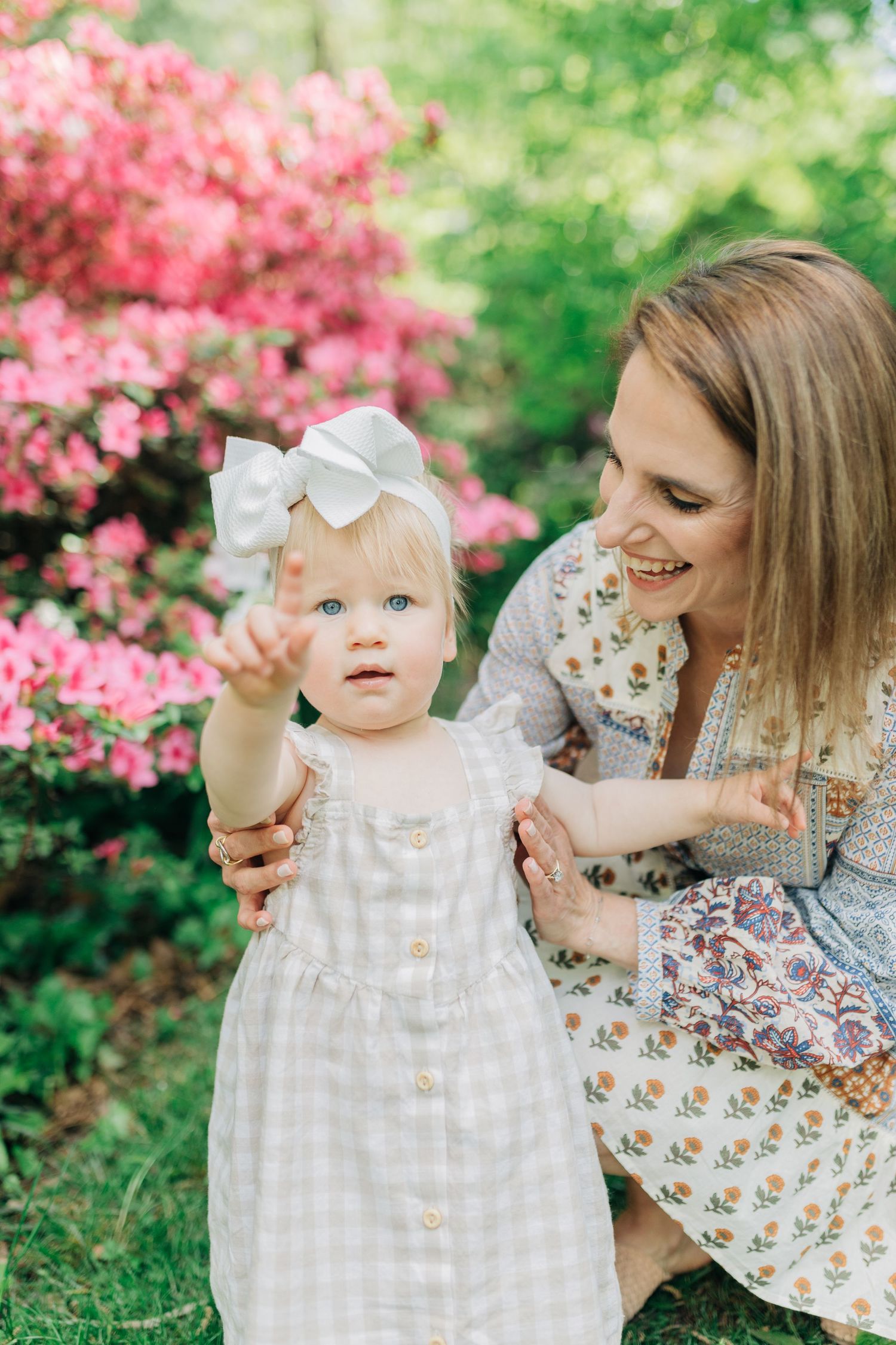 Spring portrait session with baby in white dress among blooming azalea bushes.