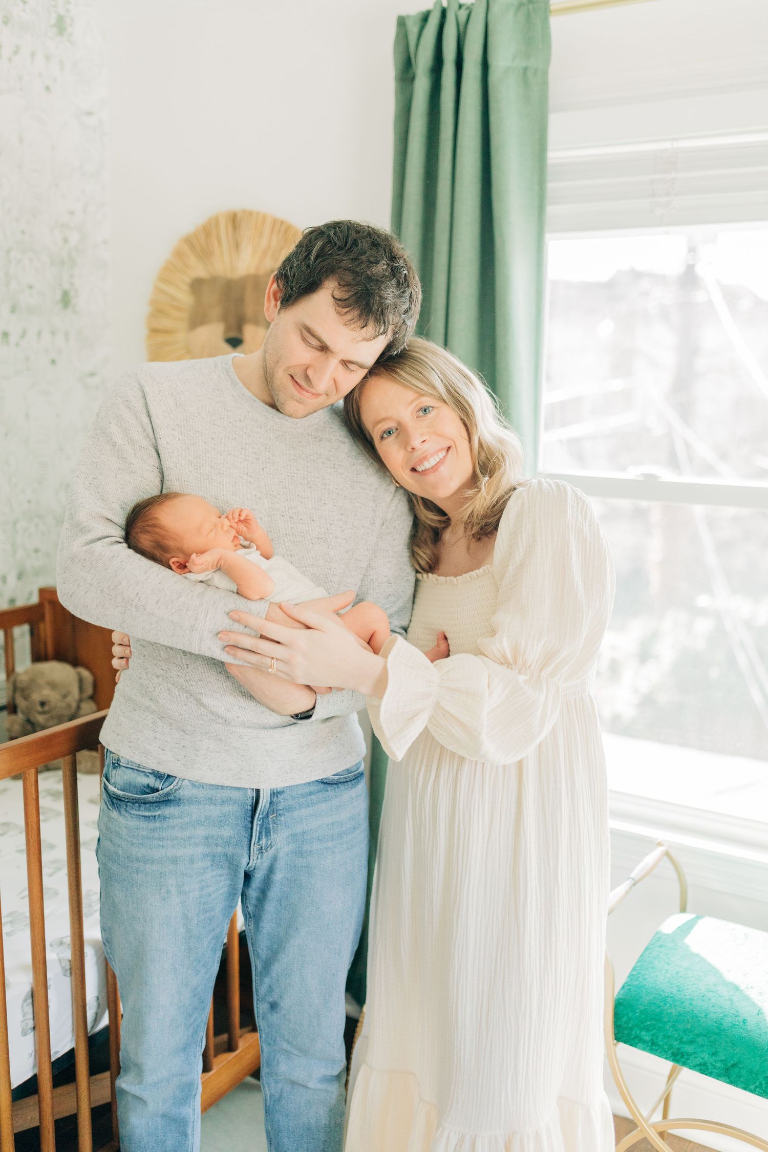 Parents in white and gray stand together holding newborn baby in nursery with green curtains.