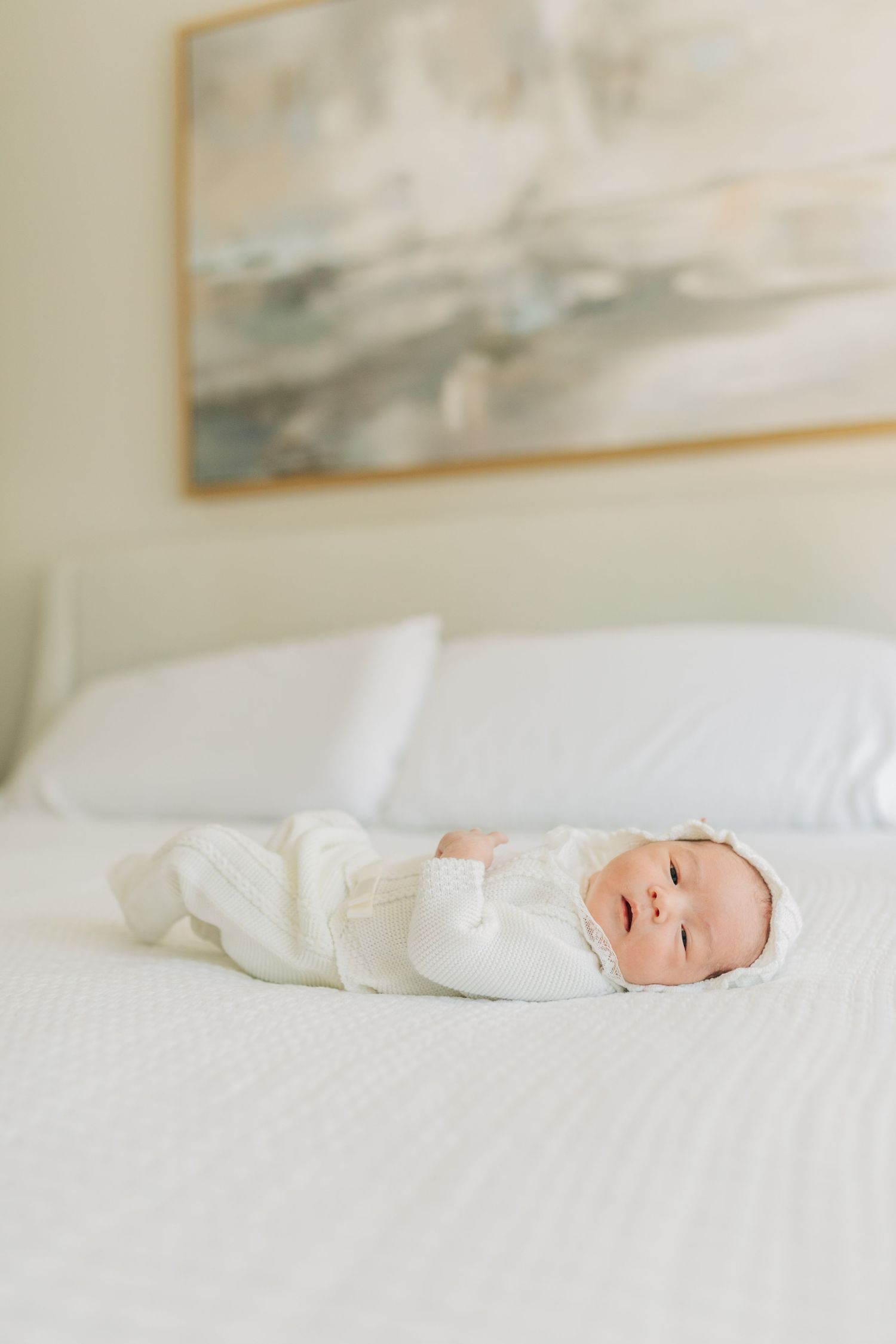 Infant in white clothing lying on bed beneath framed abstract artwork.