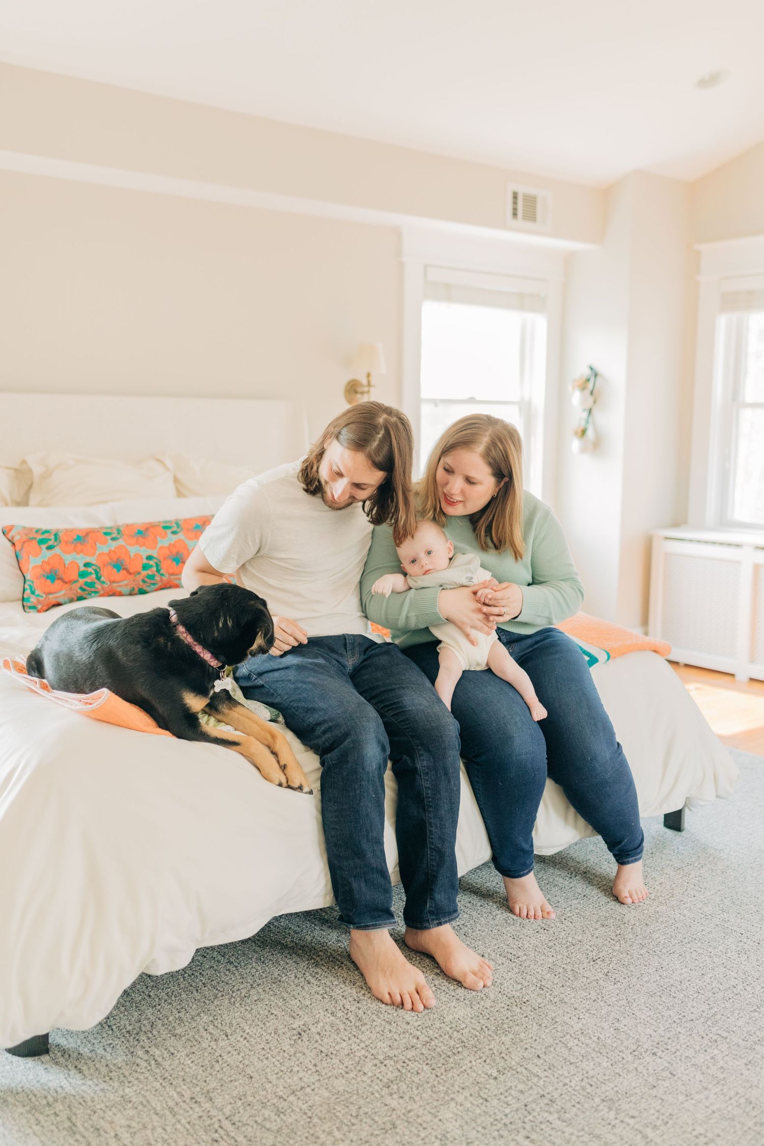 Family shares playful interactions on white bed with decorative orange pillow.