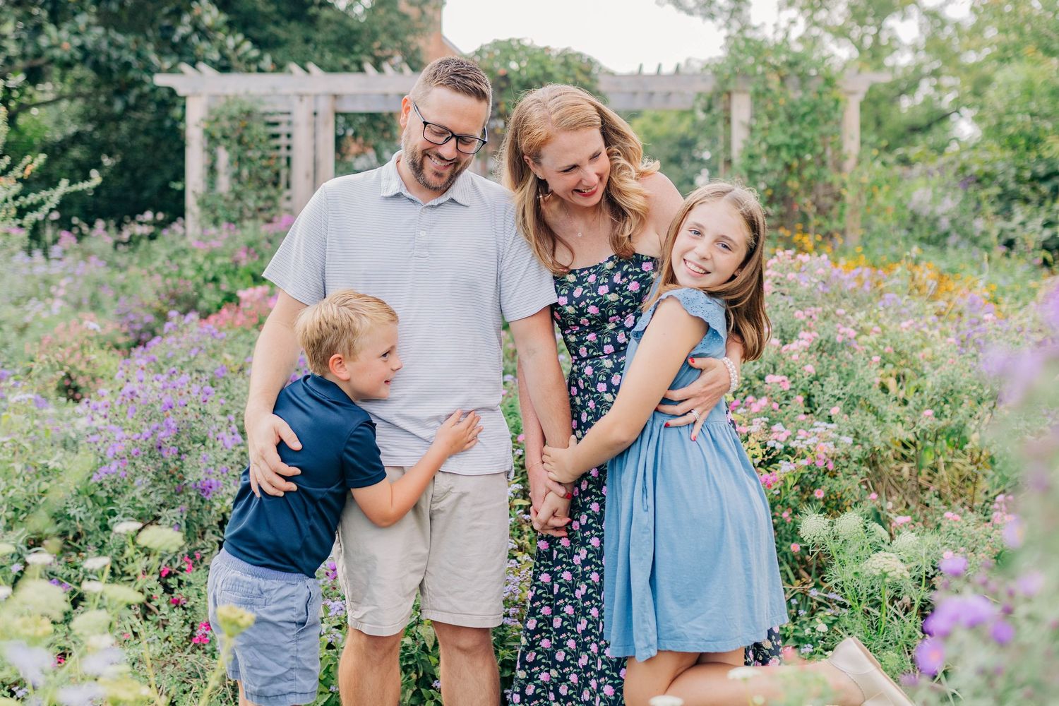A happy family embraces while standing in a lush garden with purple and yellow blooms.