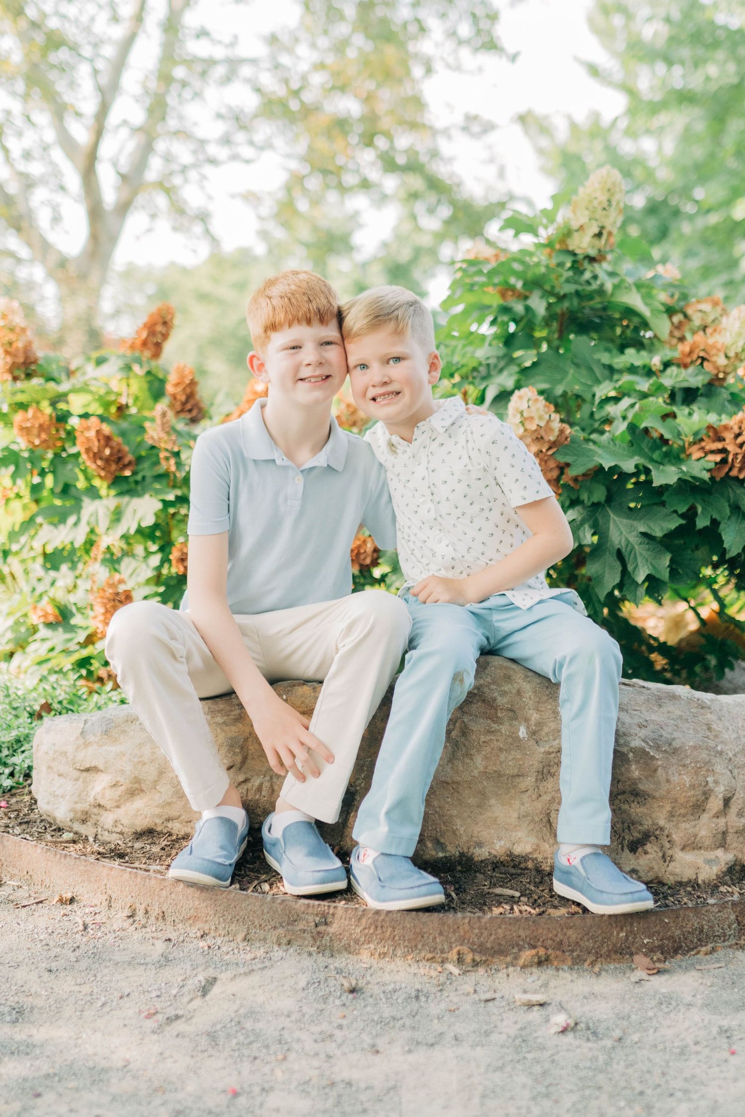 Children seated on a stone wall surrounded by green foliage and orange flowers in a natural outdoor setting.