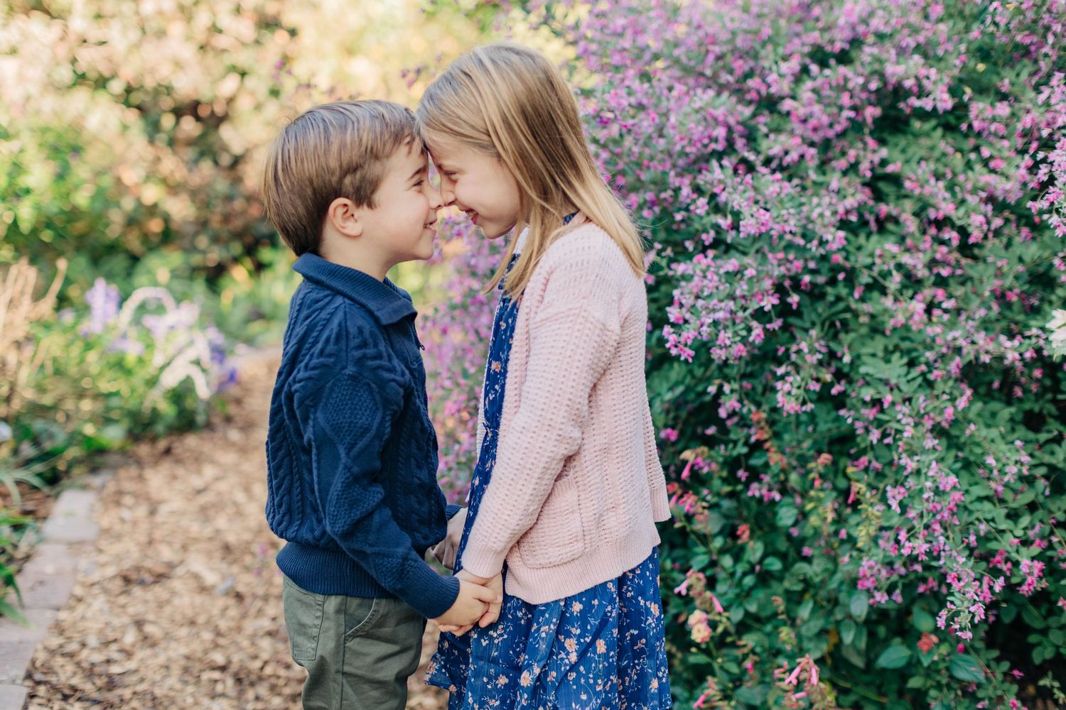 Two young children touch foreheads and smile while holding hands in a lush garden with purple flowering bushes.