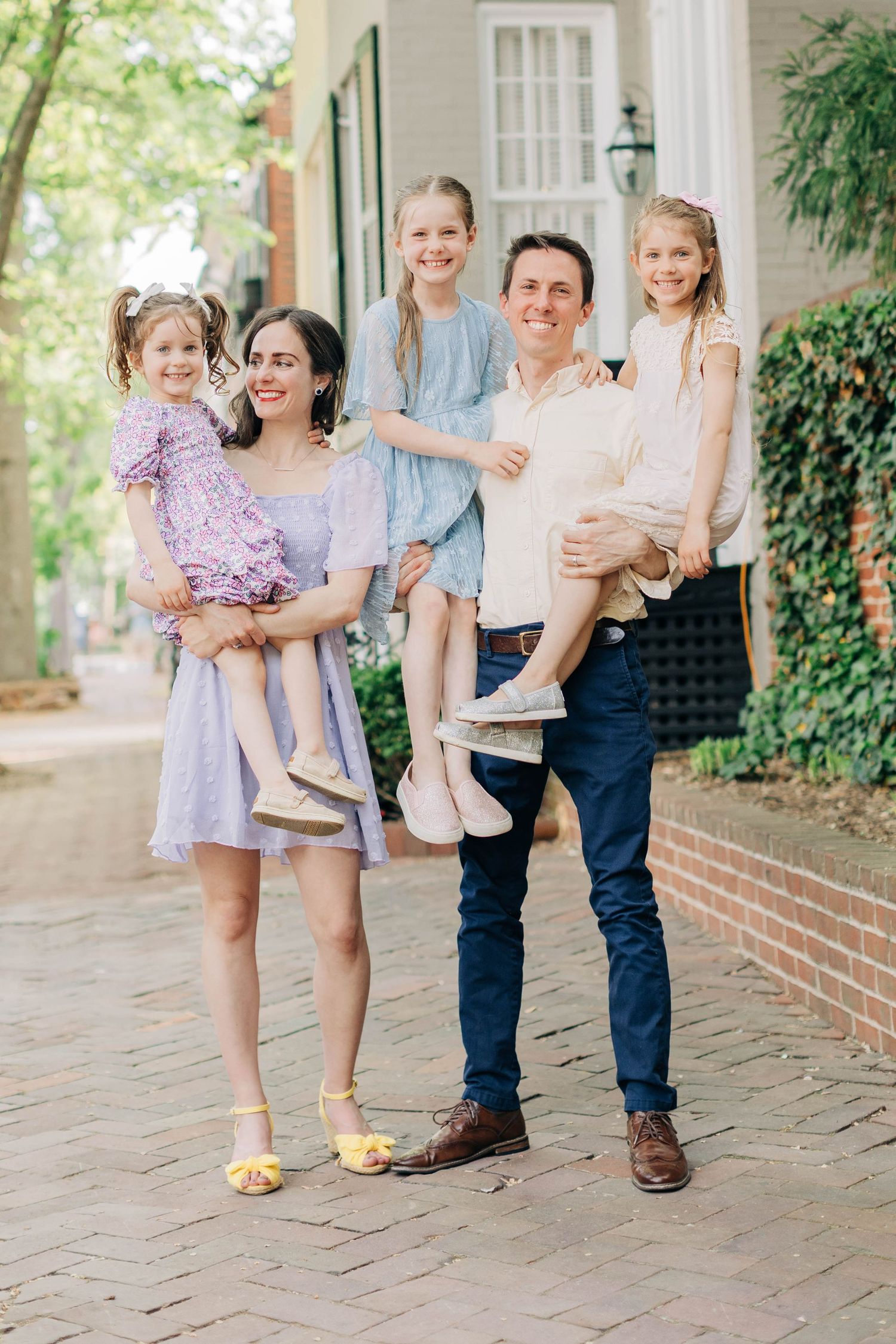 Happy family of five posing outdoors on brick path, parents holding three young daughters in pastel dresses.