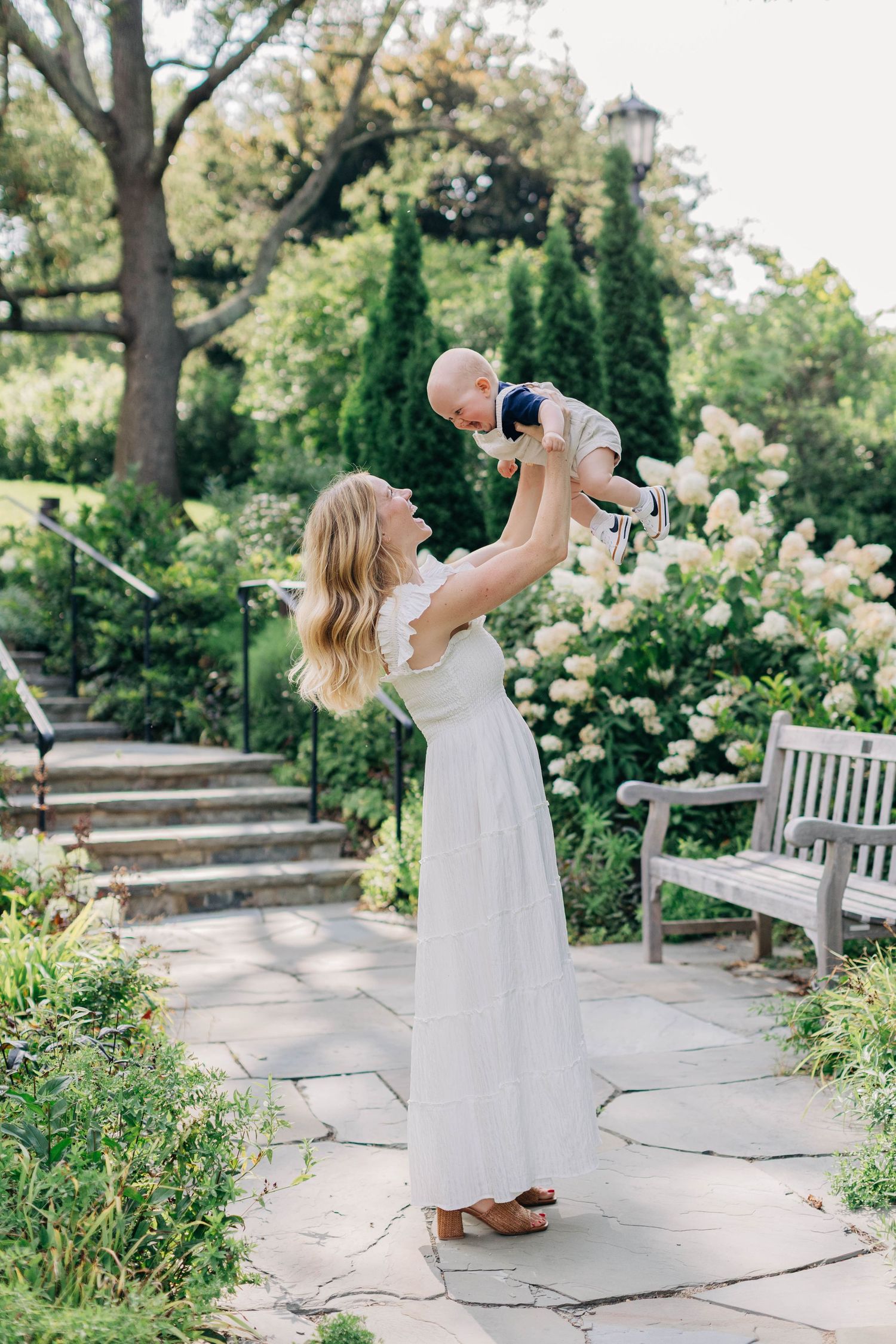 Joyful mother in white dress lifting smiling baby overhead in a beautiful garden with white hydrangeas.