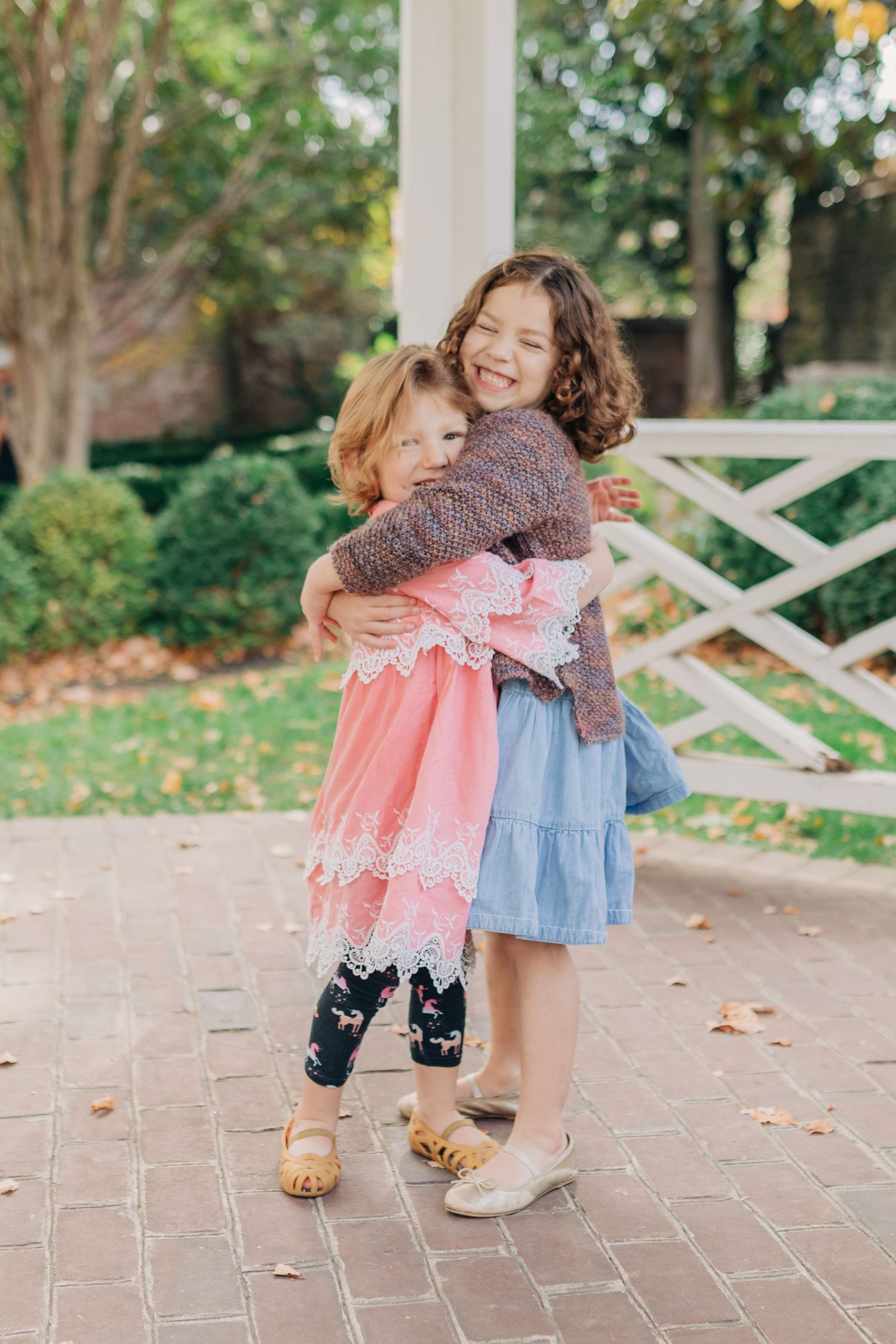 Two young sisters share a warm hug outdoors, wearing colorful dresses on a brick patio near a white fence.