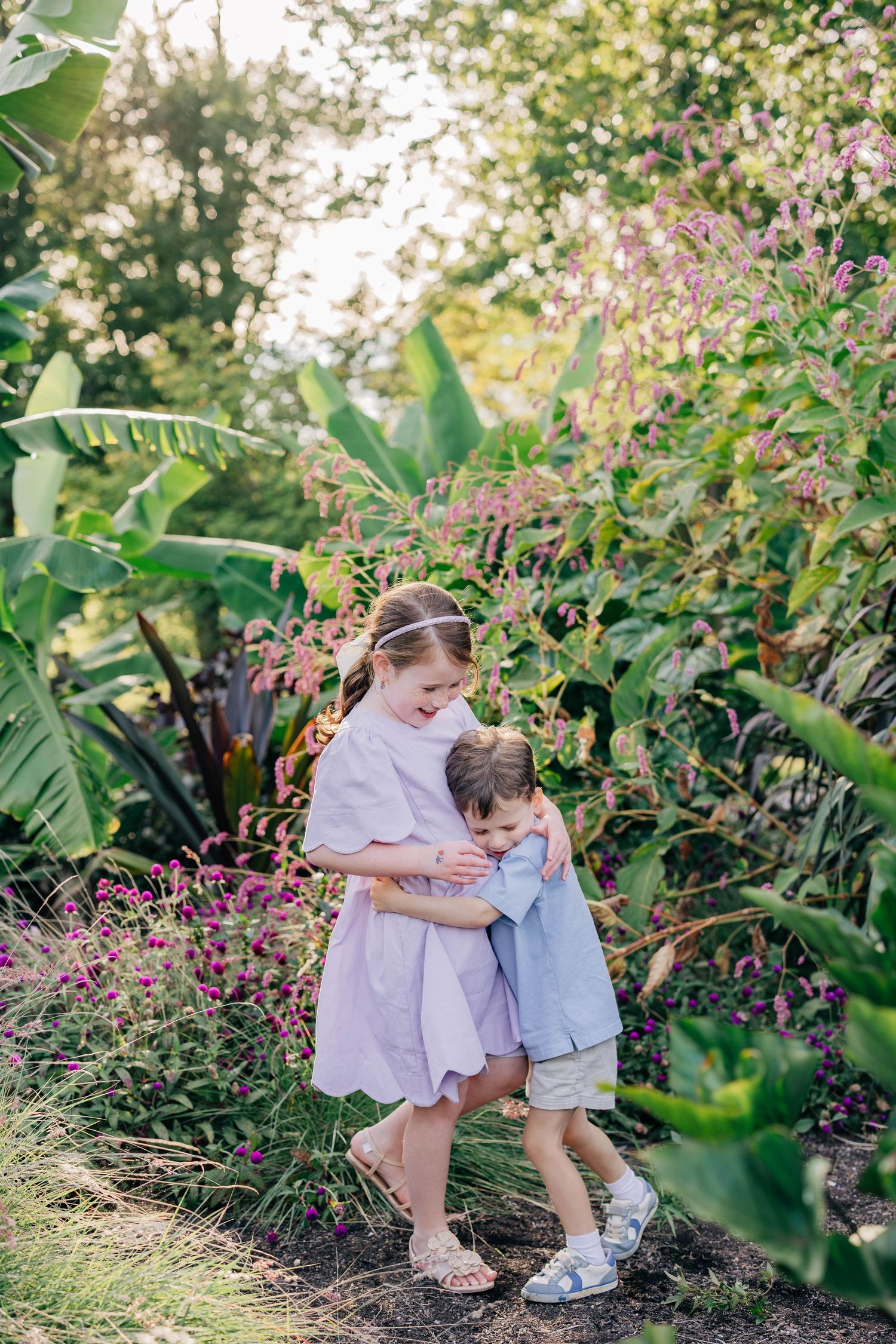 Two children hugging in a lush garden surrounded by tropical plants and purple flowers in warm sunlight.