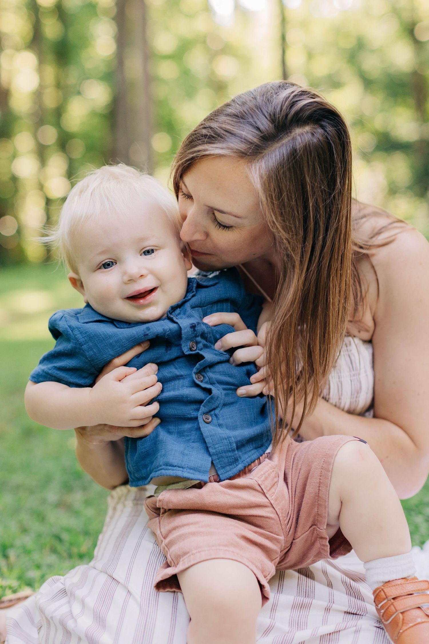 A loving mother kisses her smiling baby boy on the cheek while sitting outdoors in a lush green park.