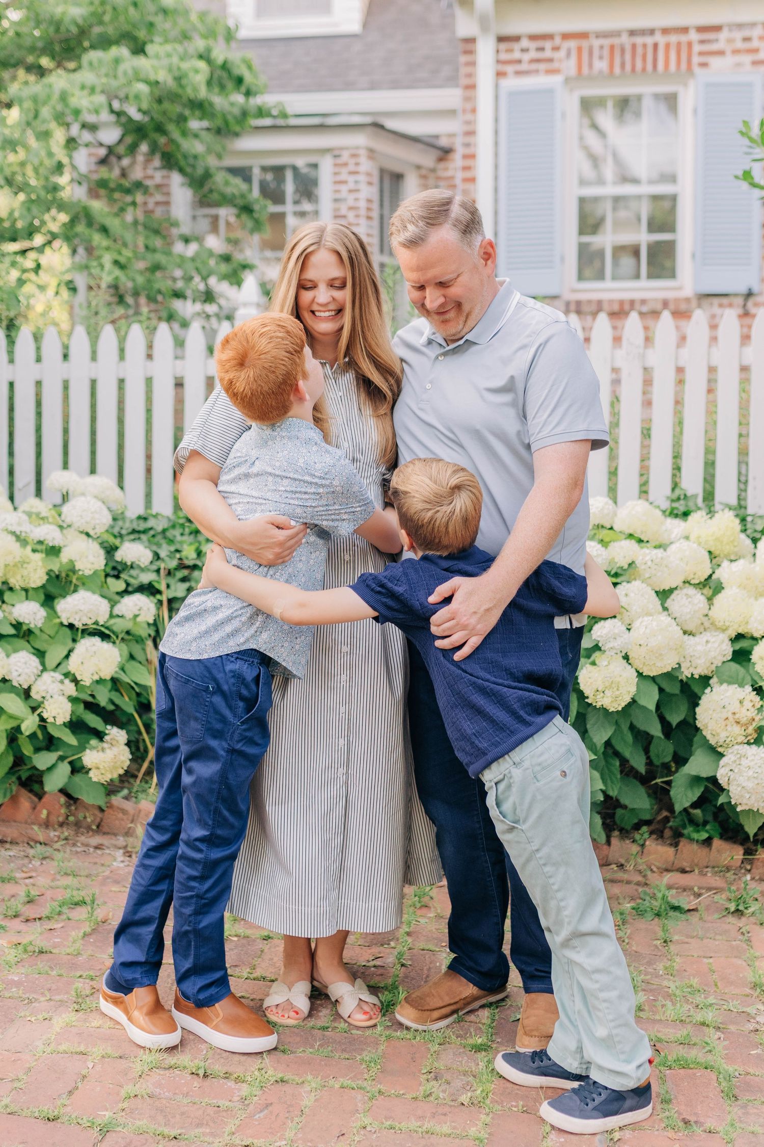 Happy family of four hugging outdoors near white picket fence with blooming hydrangeas.