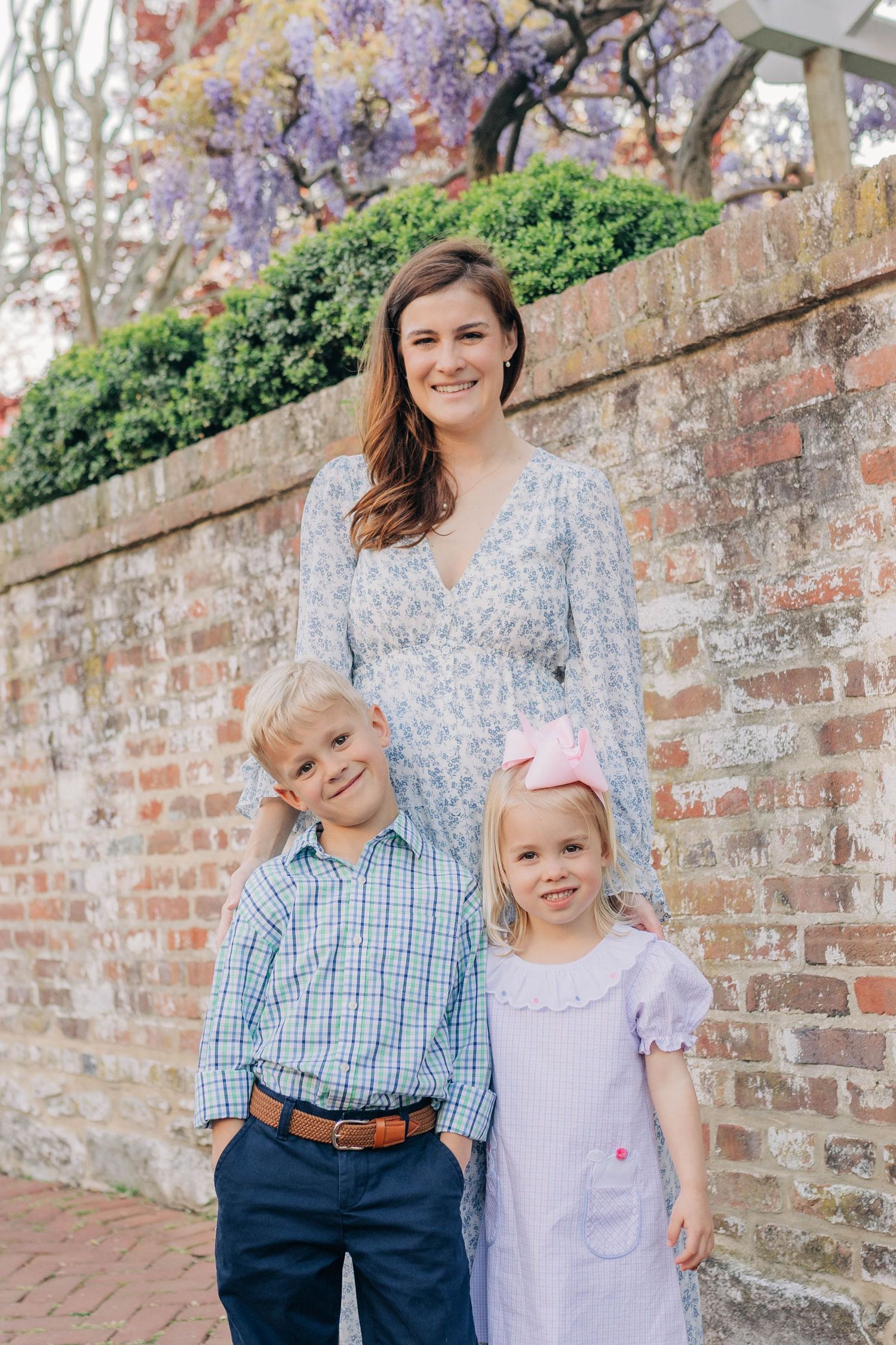 A smiling mother in a floral dress poses with her two young children against a brick wall with purple wisteria.