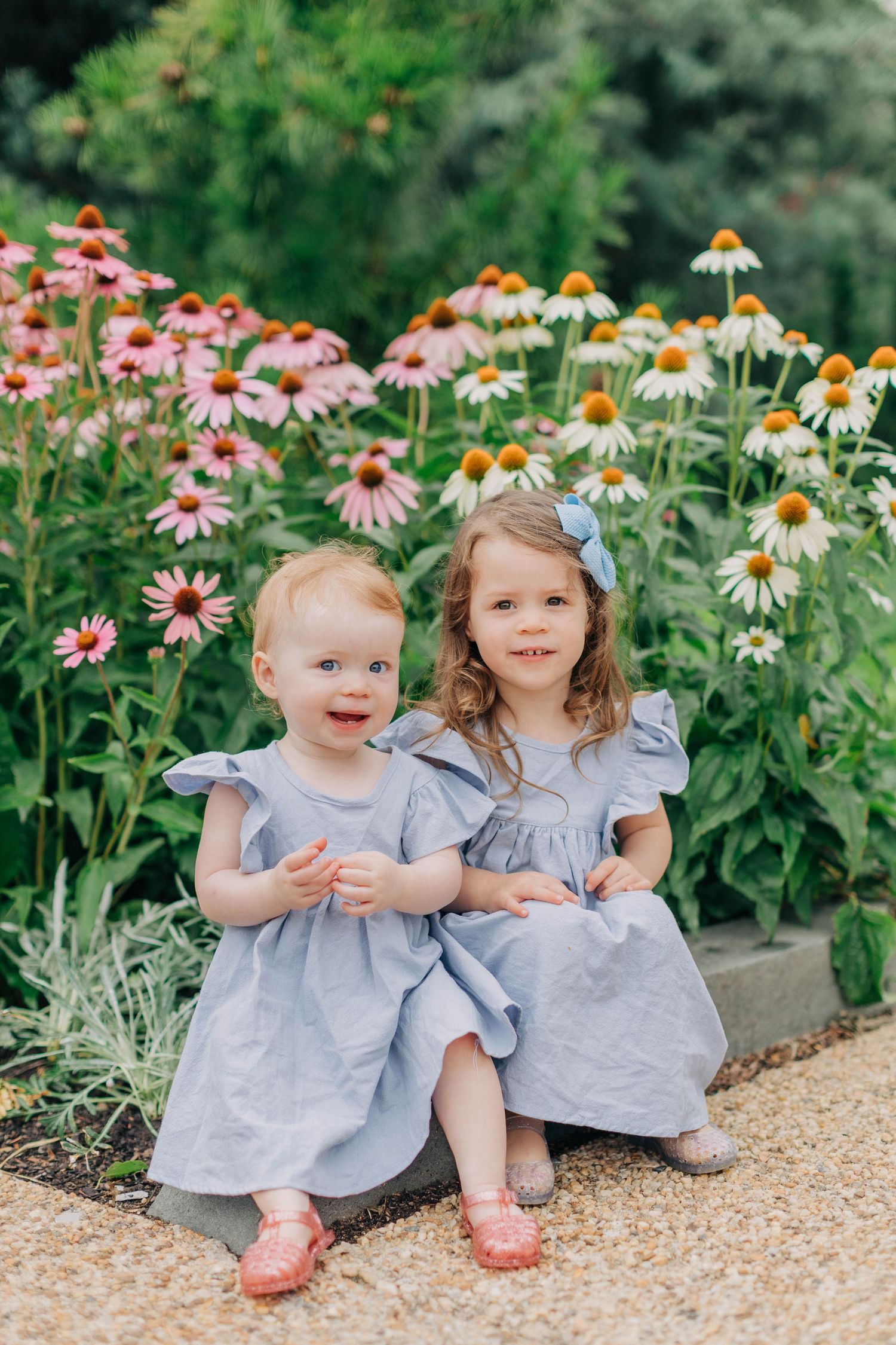 Two little girls in matching blue dresses sitting together in a garden with pink coneflowers blooming behind them.