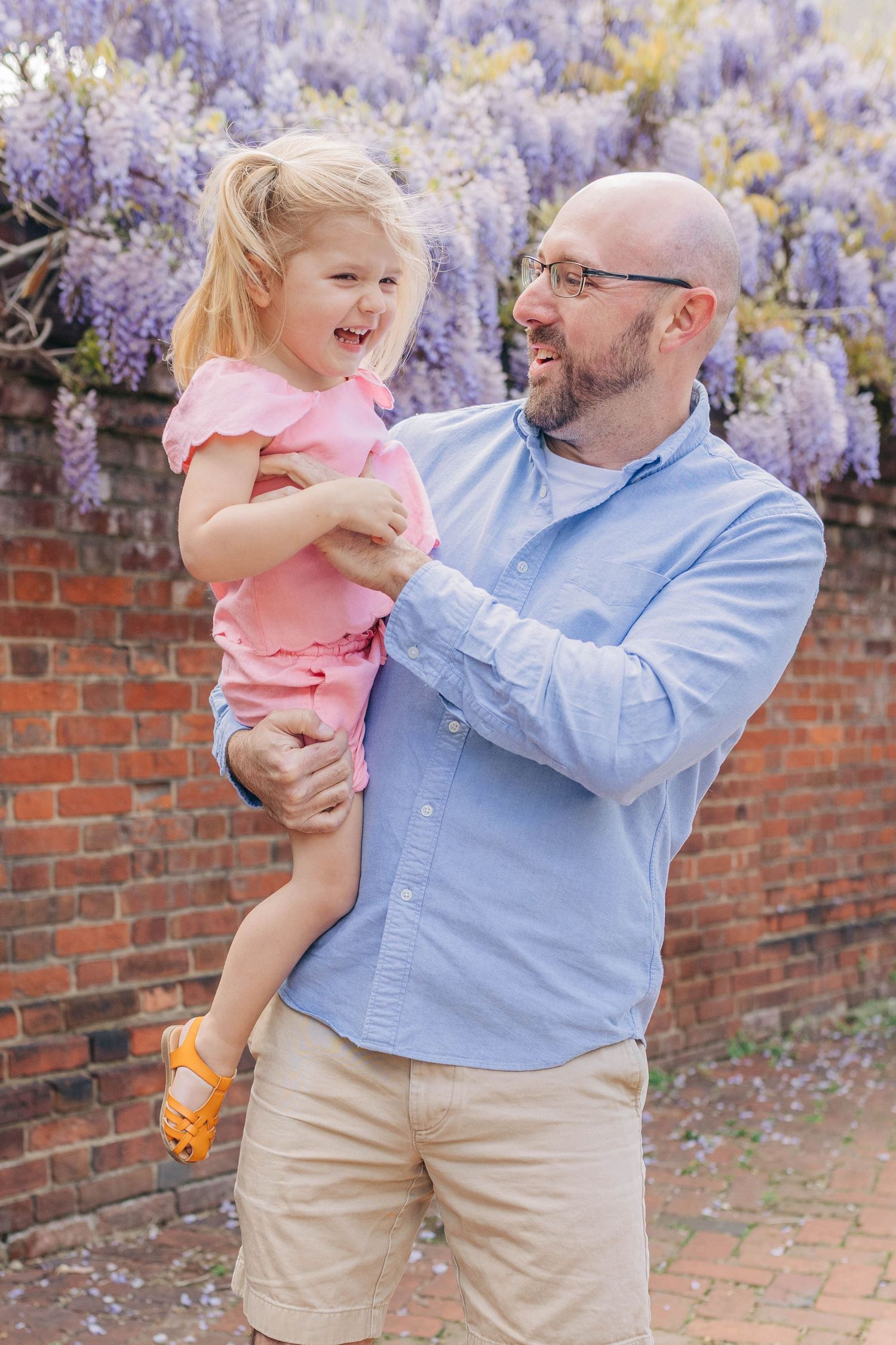 Smiling bald father holding laughing blonde girl in pink dress near wisteria-covered brick wall outdoors.
