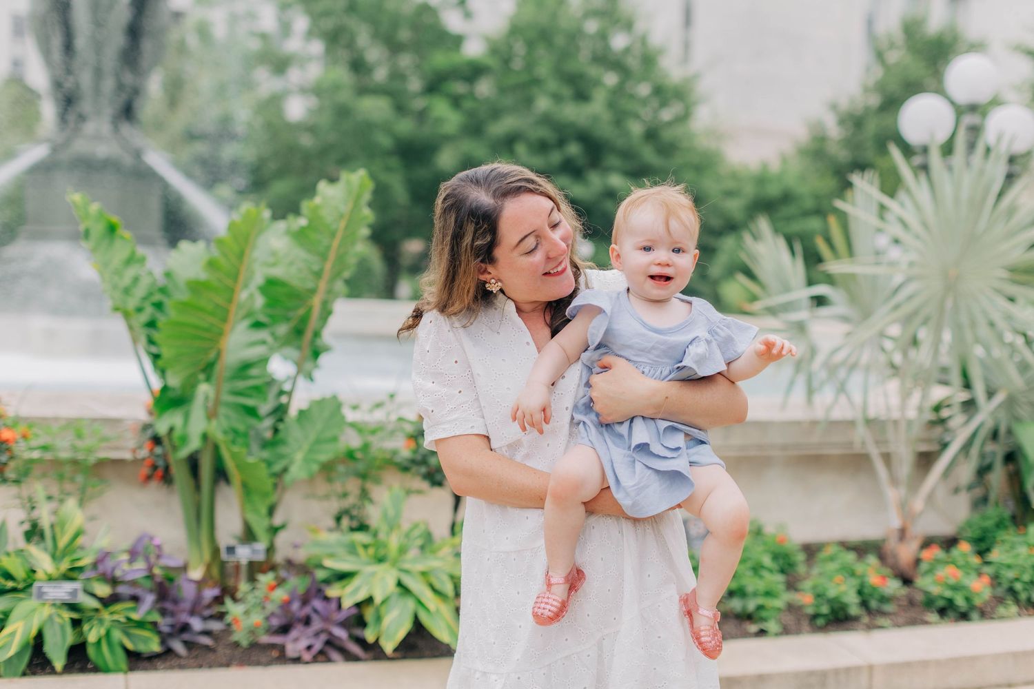Smiling mother holding her happy baby girl in a blue dress in front of a garden fountain.
