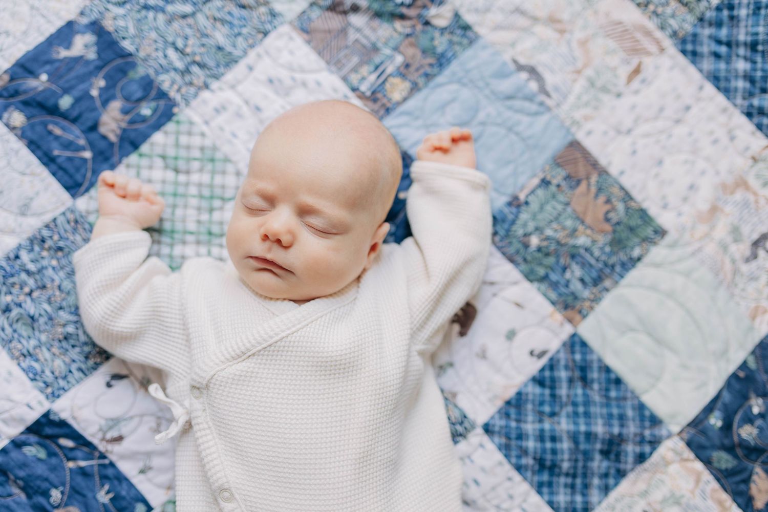 Sleeping newborn baby in white onesie lying on a blue and white patchwork quilt with arms raised.