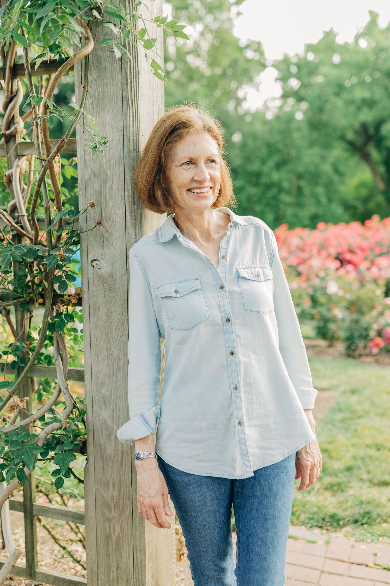 Person in light blue shirt smiles warmly while standing near wooden garden trellis.