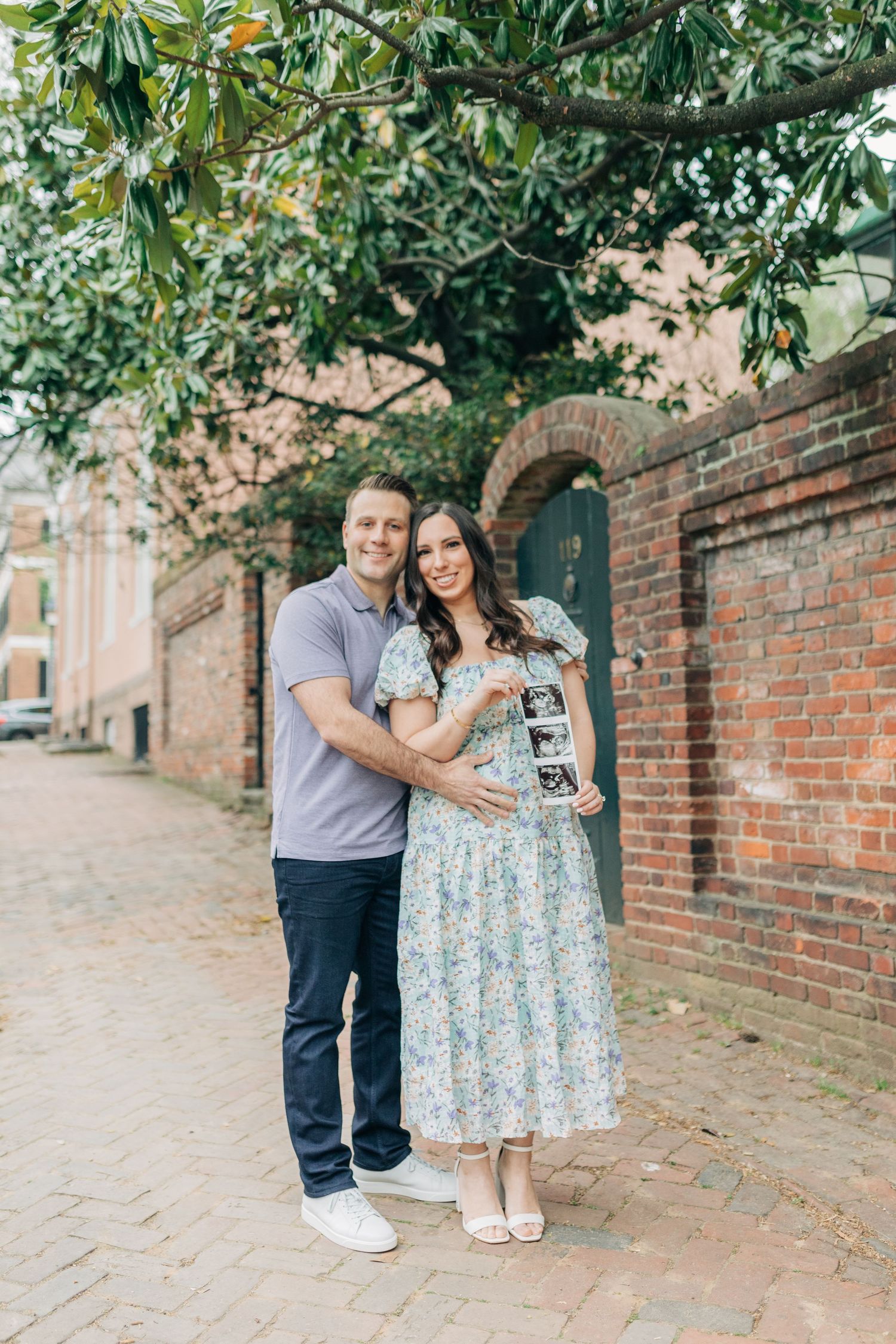 A couple poses with ultrasound photos along a tree-lined brick pathway.