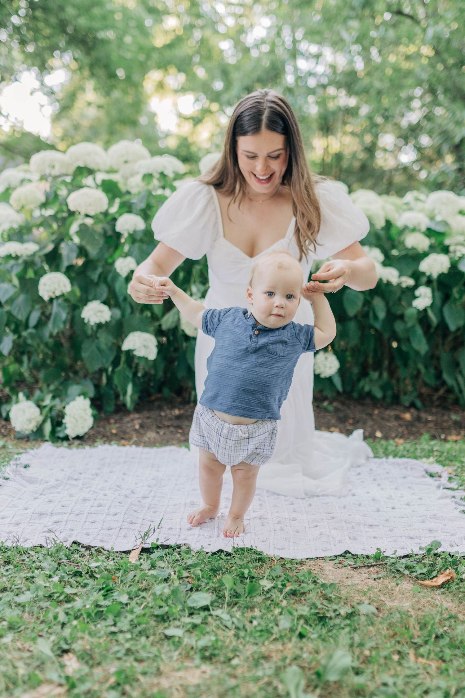 Playful moments captured on white blanket with baby in denim outfit against floral garden backdrop.