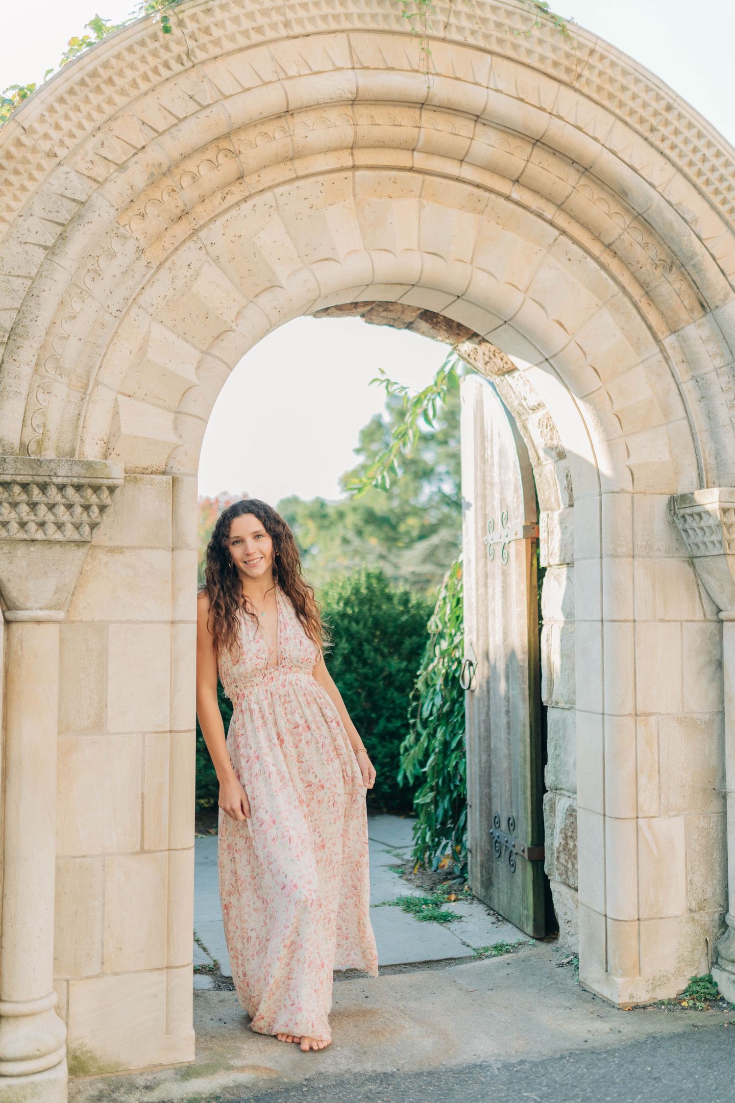 A person in a floral dress poses beneath stone archways in a garden setting.