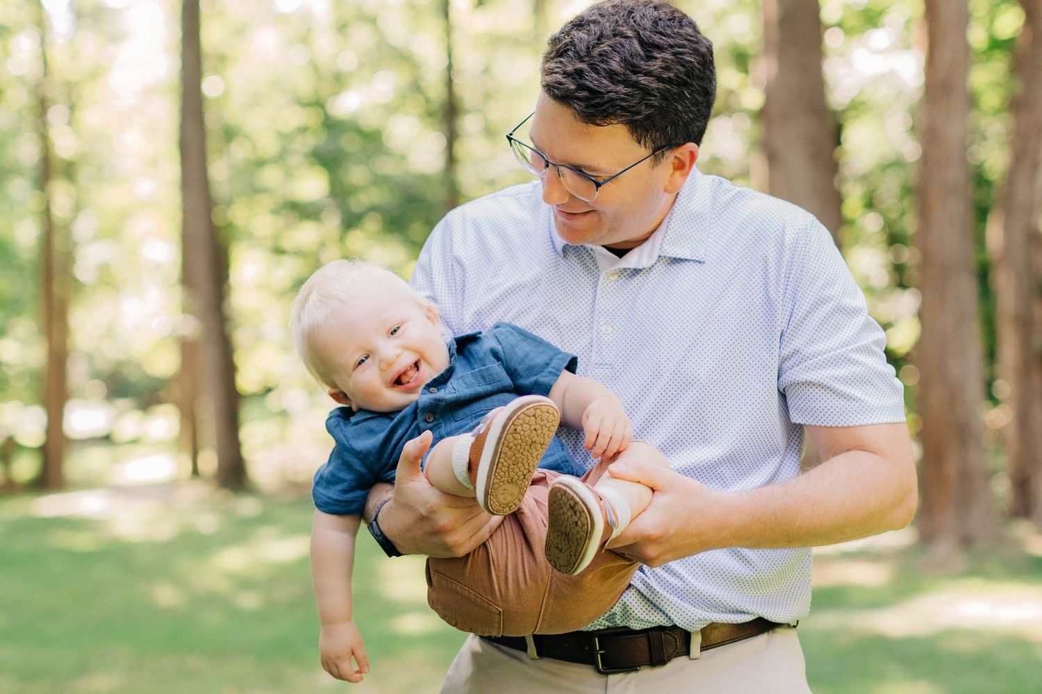 A smiling father holds his laughing baby outdoors in a lush green park setting.