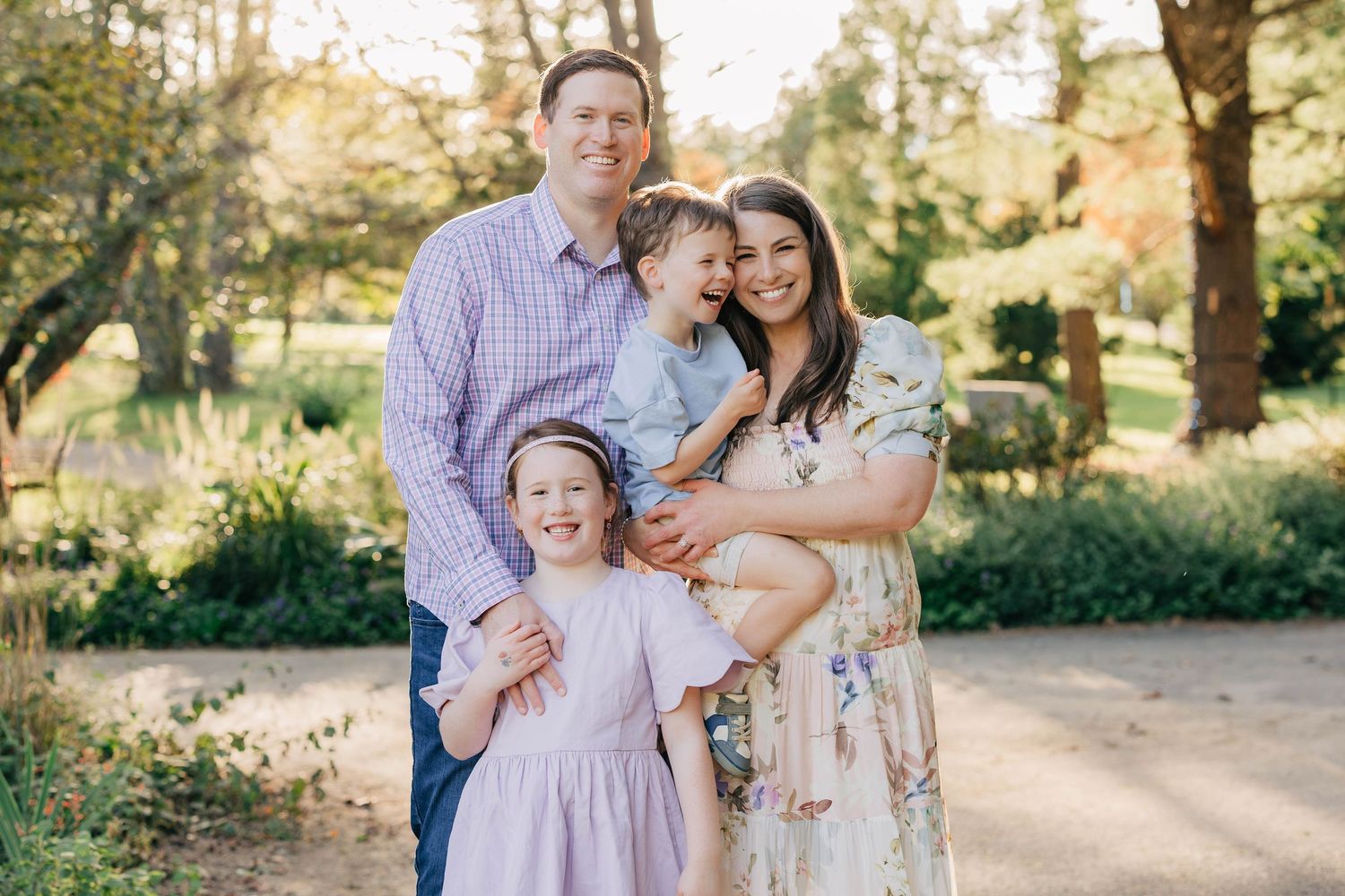 Happy family of four posing outdoors in a lush garden, parents smiling with two young children in spring attire.