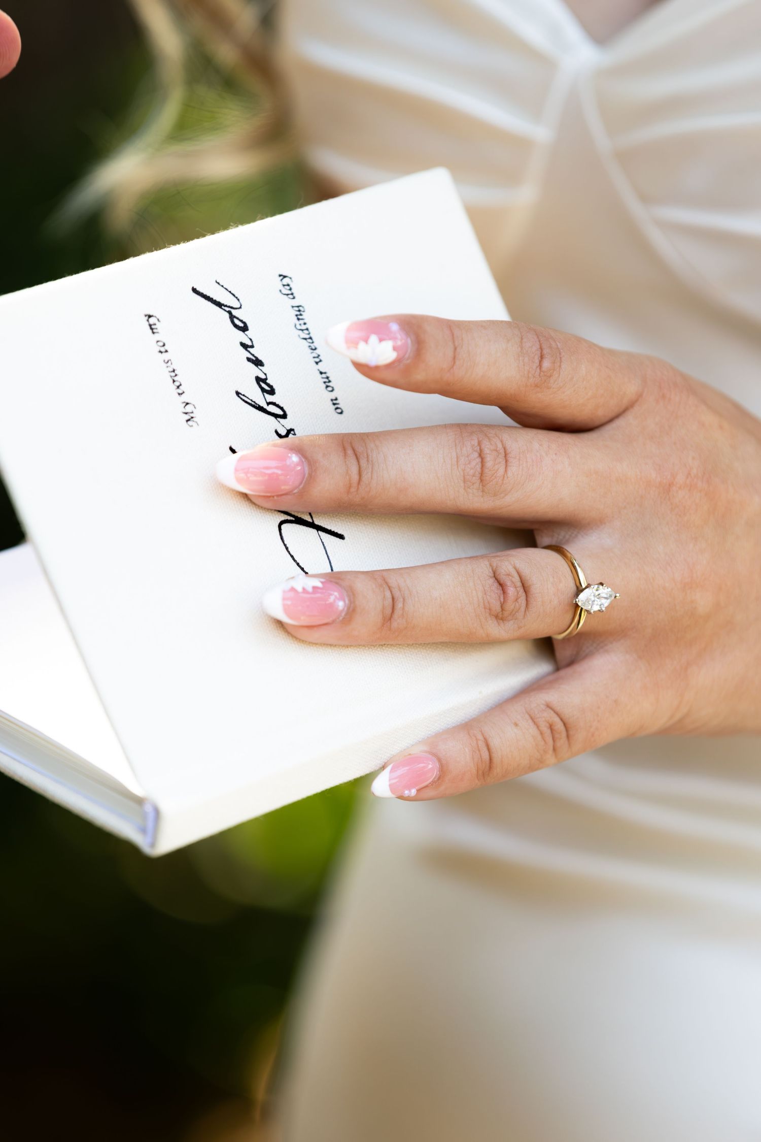 Bride holding a wedding book with pink manicured nails and a gold diamond engagement ring on her finger.