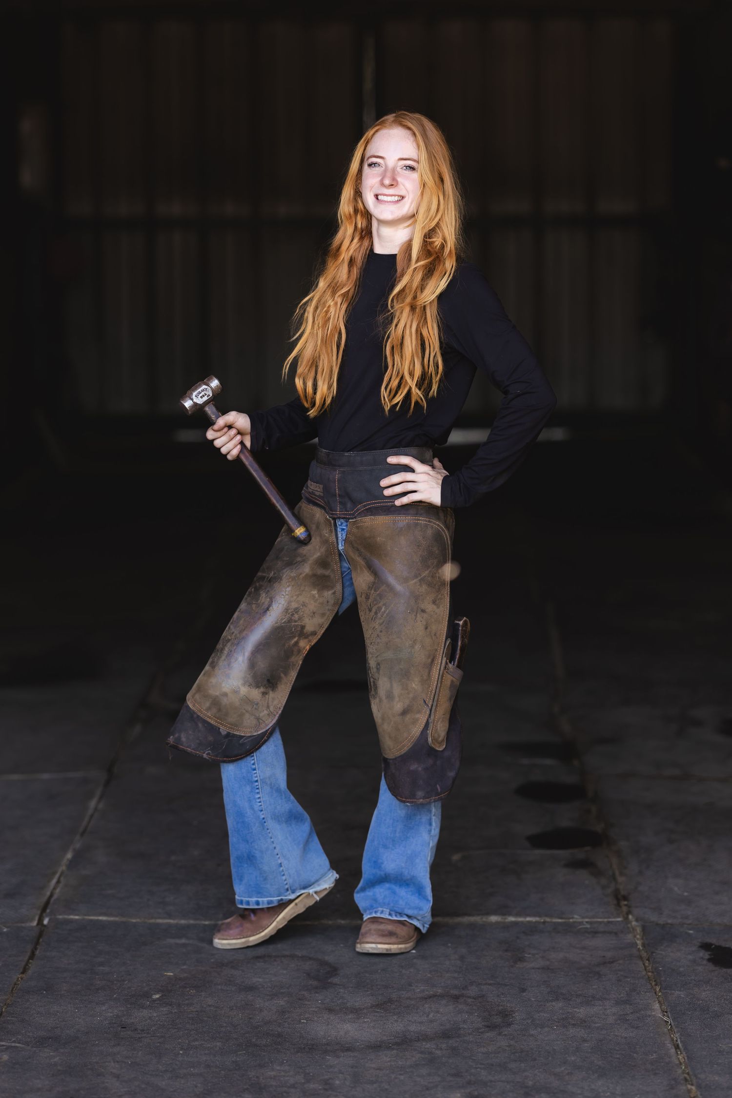 Young redhead woman in leather chaps holding a mallet, smiling confidently in a dark barn setting.