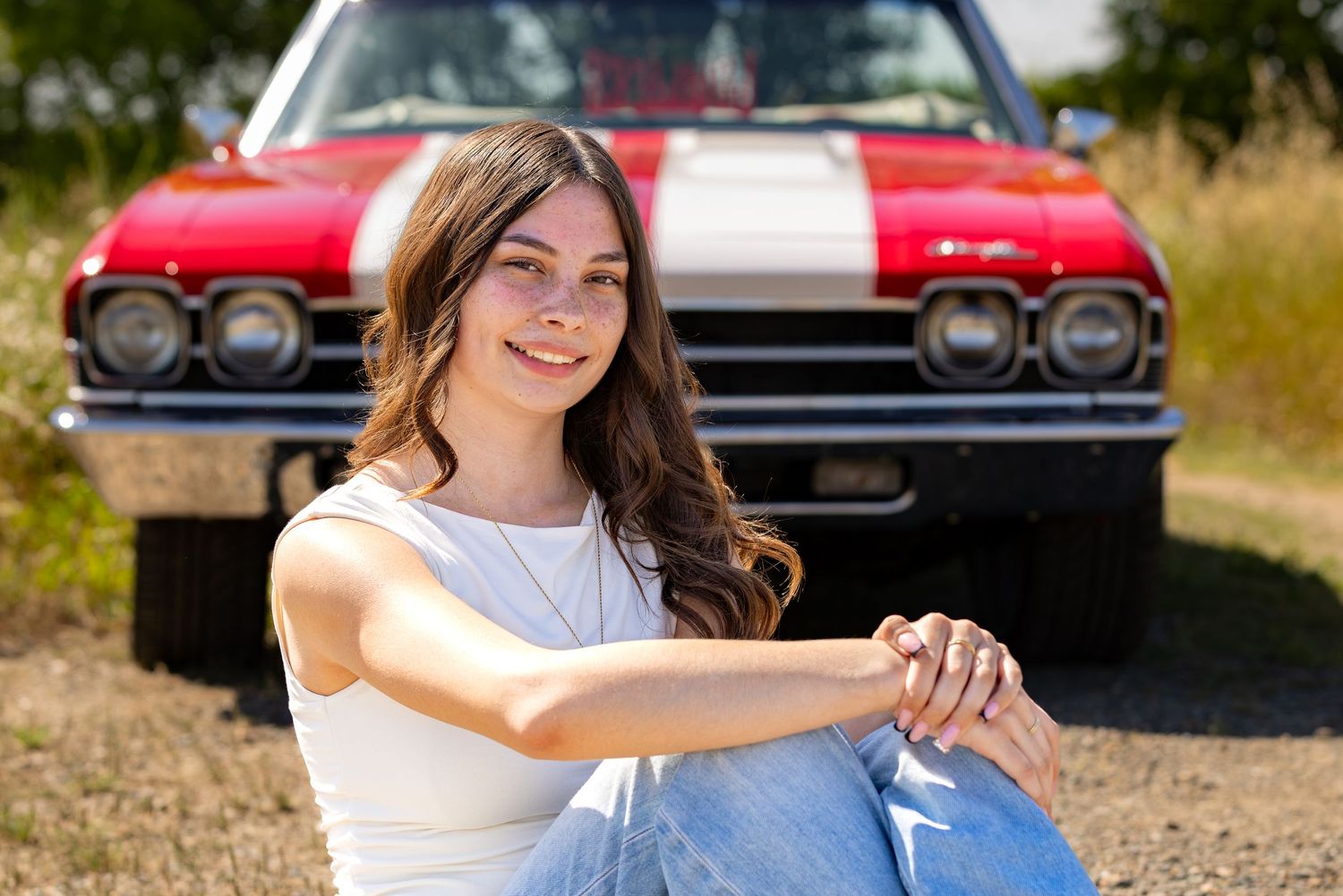 Young woman with long brown hair sits smiling in front of a classic red muscle car on a sunny day.