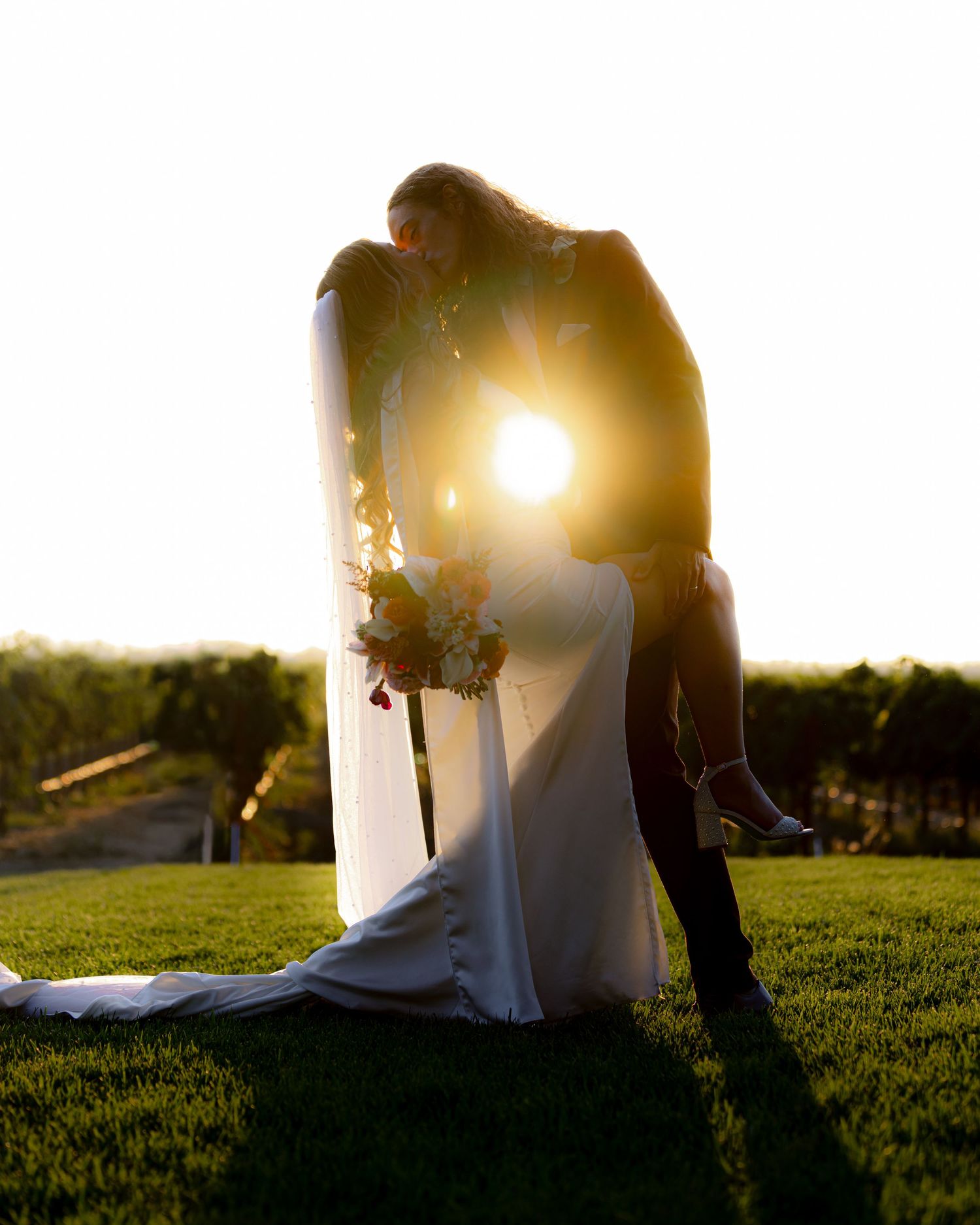 Bride and groom silhouetted against golden sunset light, sharing a kiss on green lawn with bridal bouquet and veil.