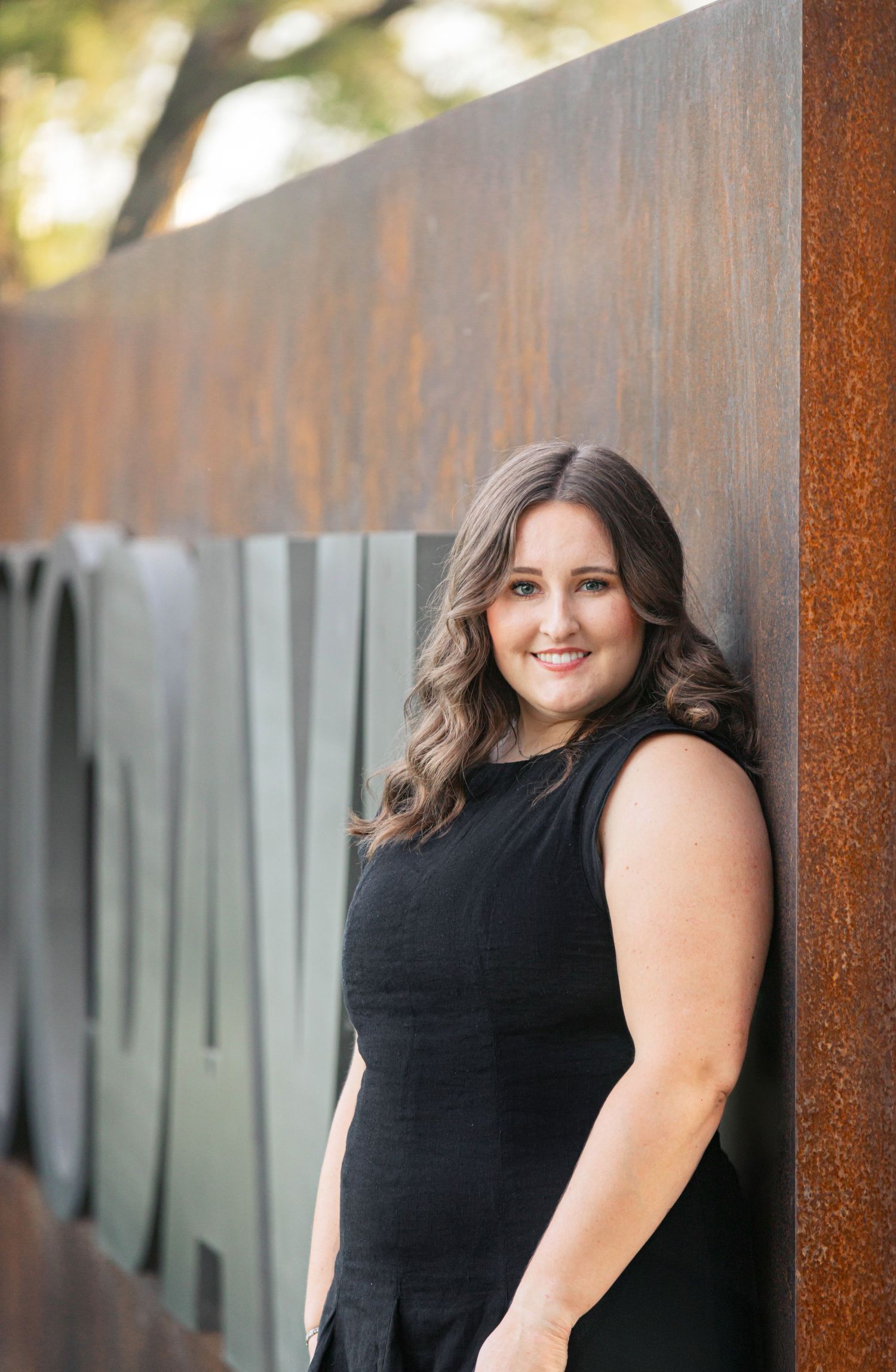 A smiling woman in a black sleeveless dress leans against a rustic metal wall outdoors in warm evening light.
