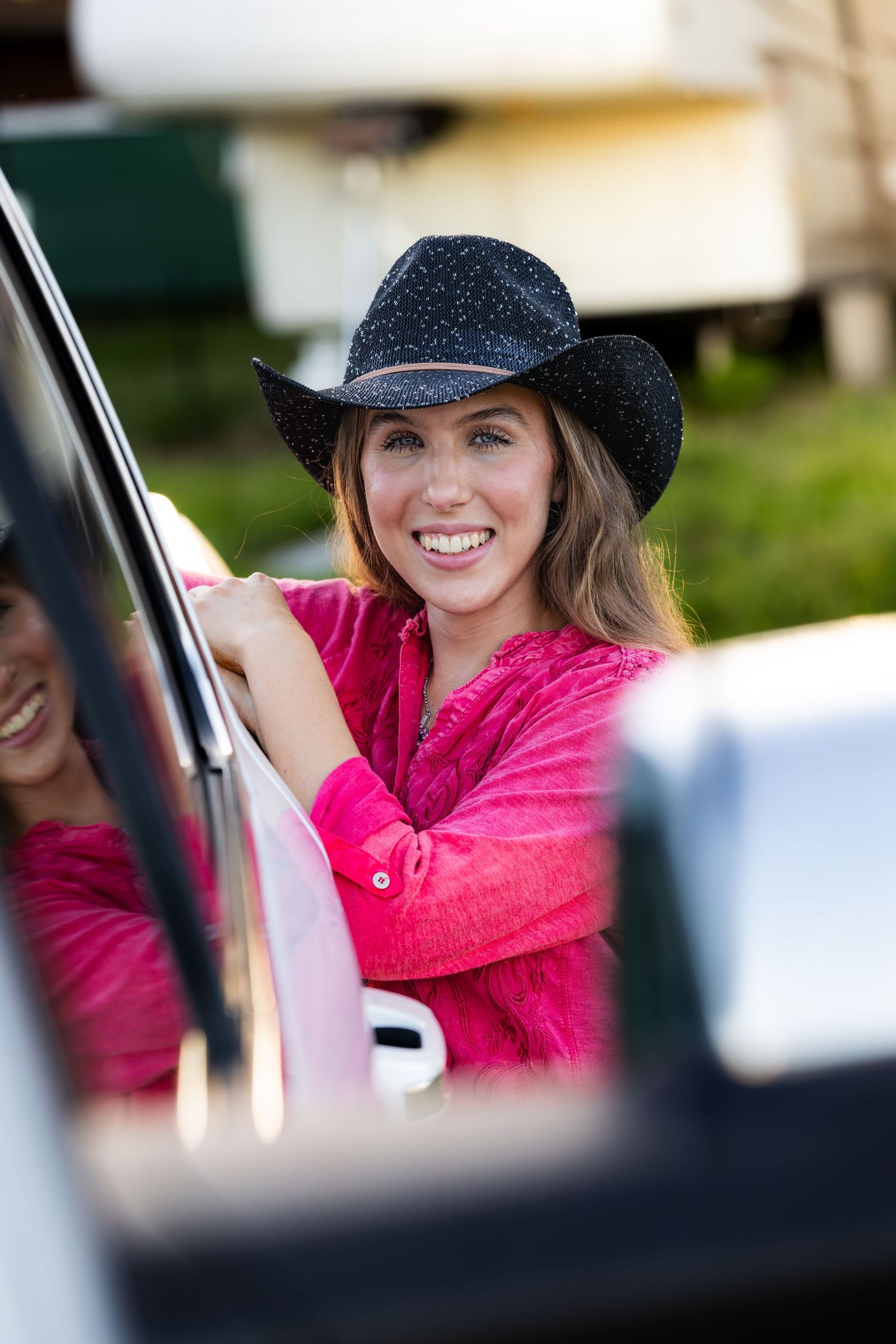 Smiling young woman in black cowboy hat and pink shirt leaning on a white truck door outdoors.