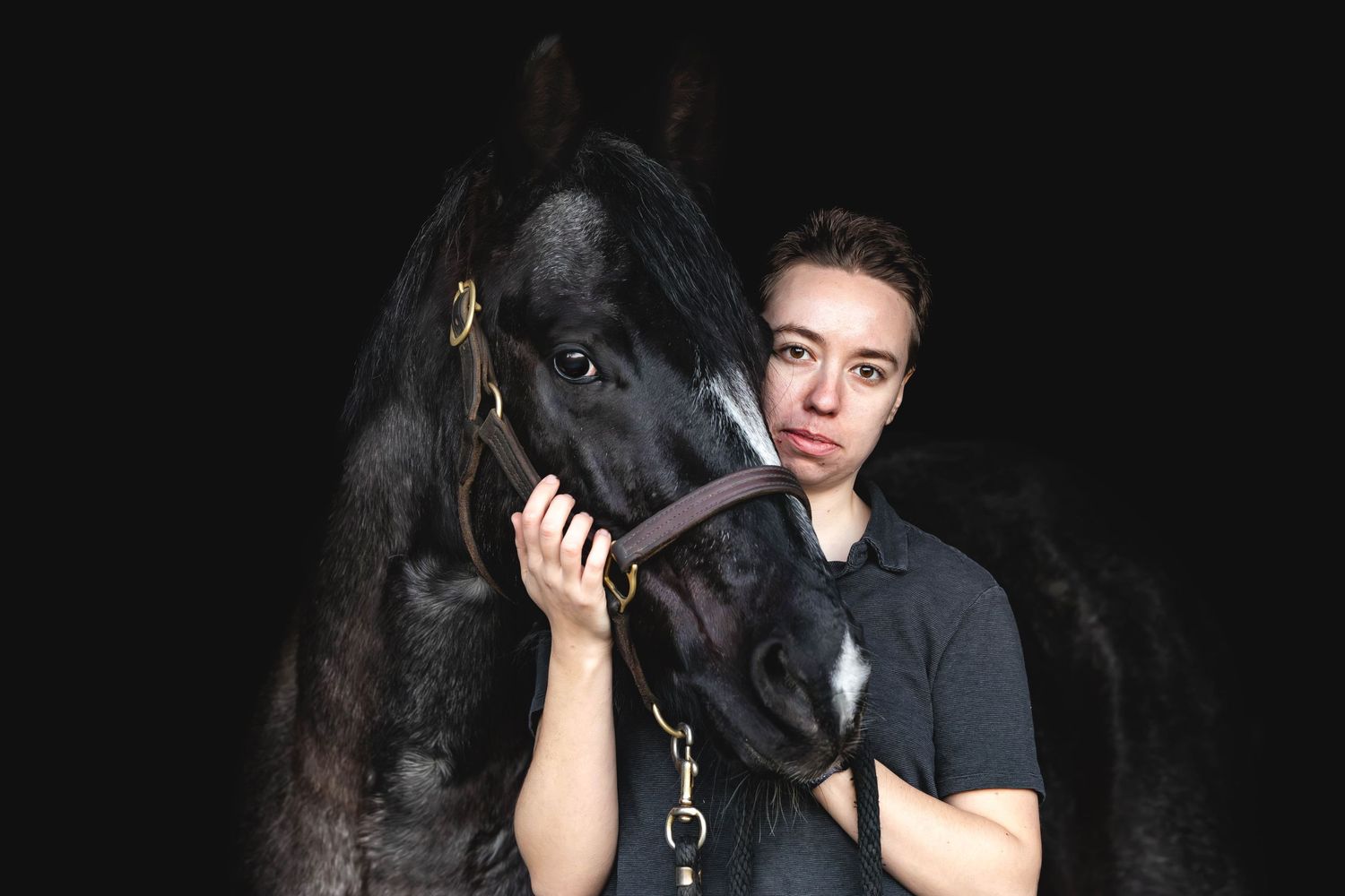 Young woman in black shirt poses closely with a dark black horse against a black background.