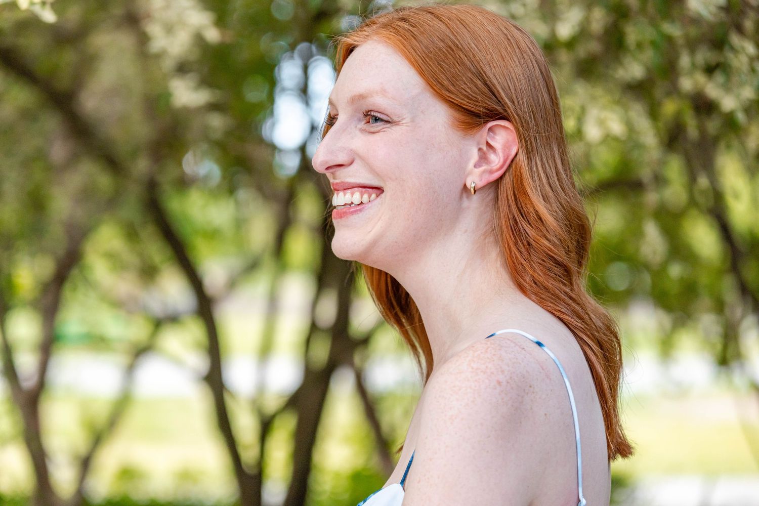 Joyful redhead woman smiling in profile outdoors with green trees in background.